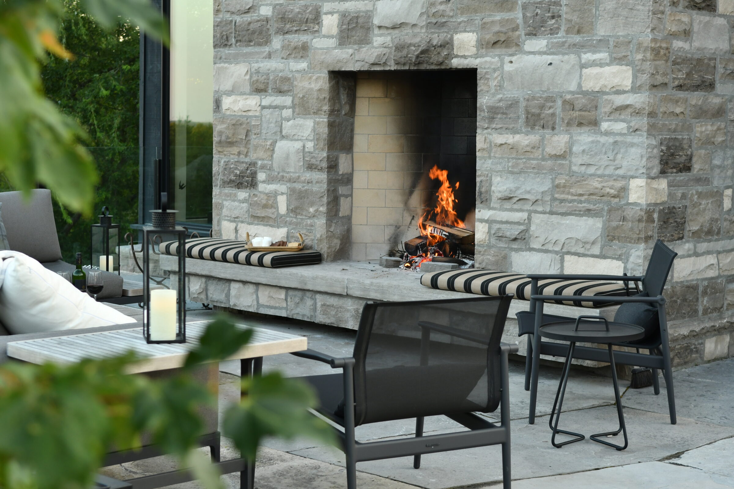 Outdoor patio featuring a cozy, stone fireplace with burning logs, surrounded by striped cushions and modern black chairs amidst a serene, leafy backdrop.