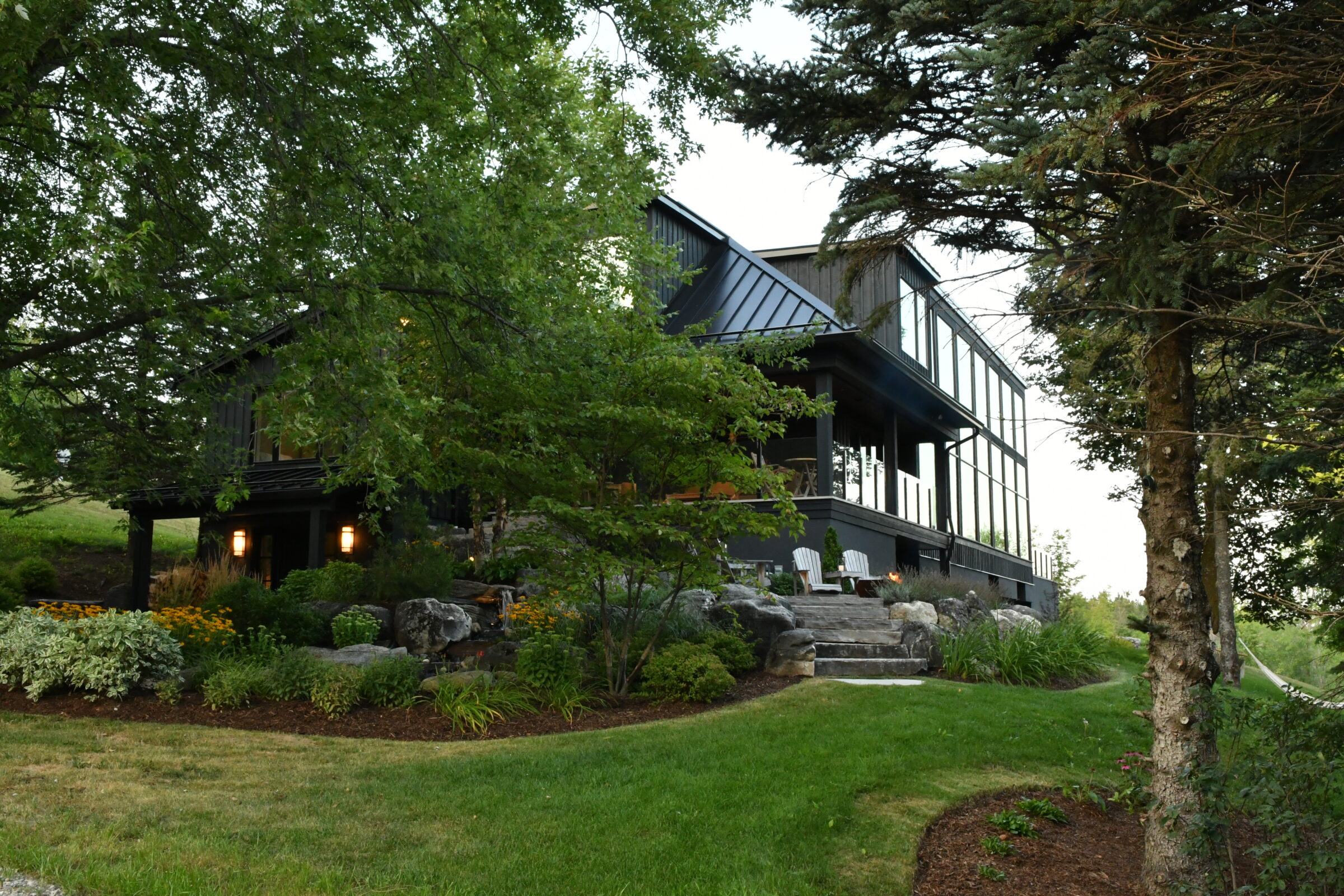 Modern house surrounded by lush greenery, featuring large windows and outdoor seating area. Well-maintained garden with diverse plants and trees.