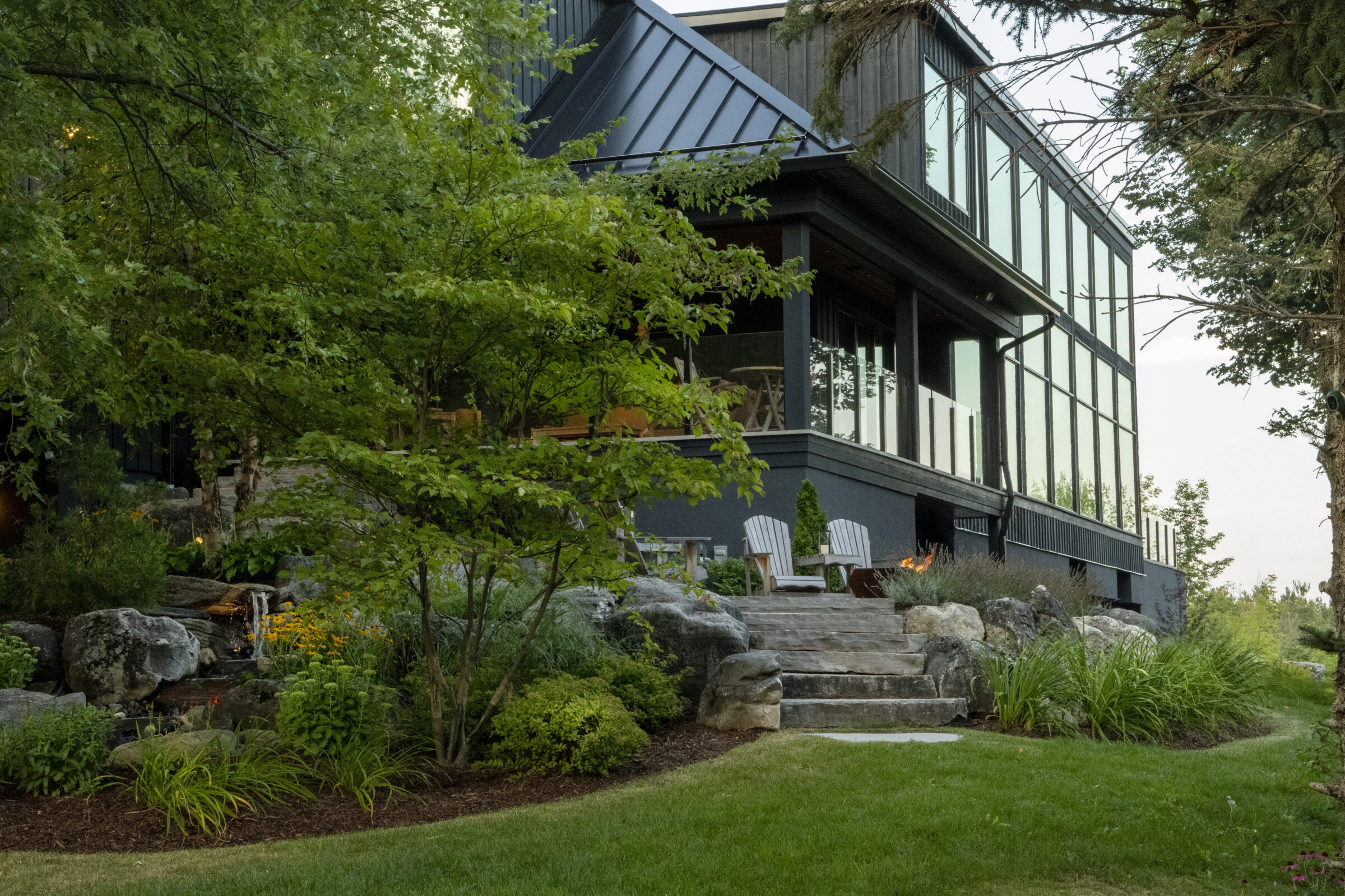 Modern house with large windows, surrounded by lush greenery. Stone pathway with chairs leads to a cozy outdoor area. Peaceful and inviting atmosphere.
