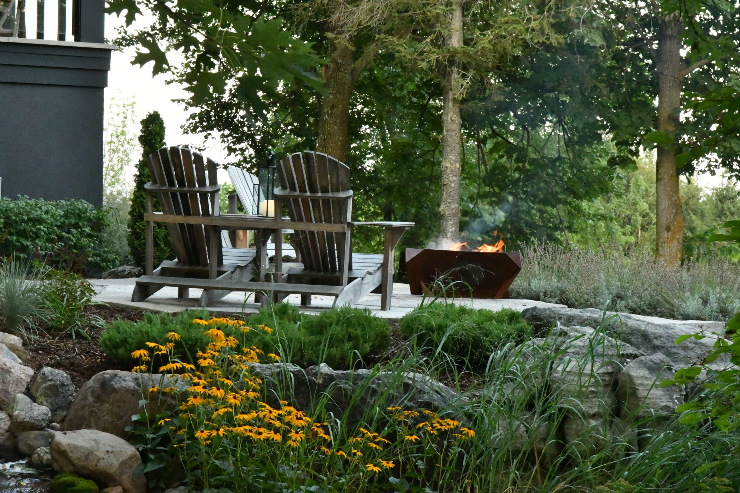Two wooden chairs face a lit fire pit amidst a lush garden with yellow flowers and surrounding trees.