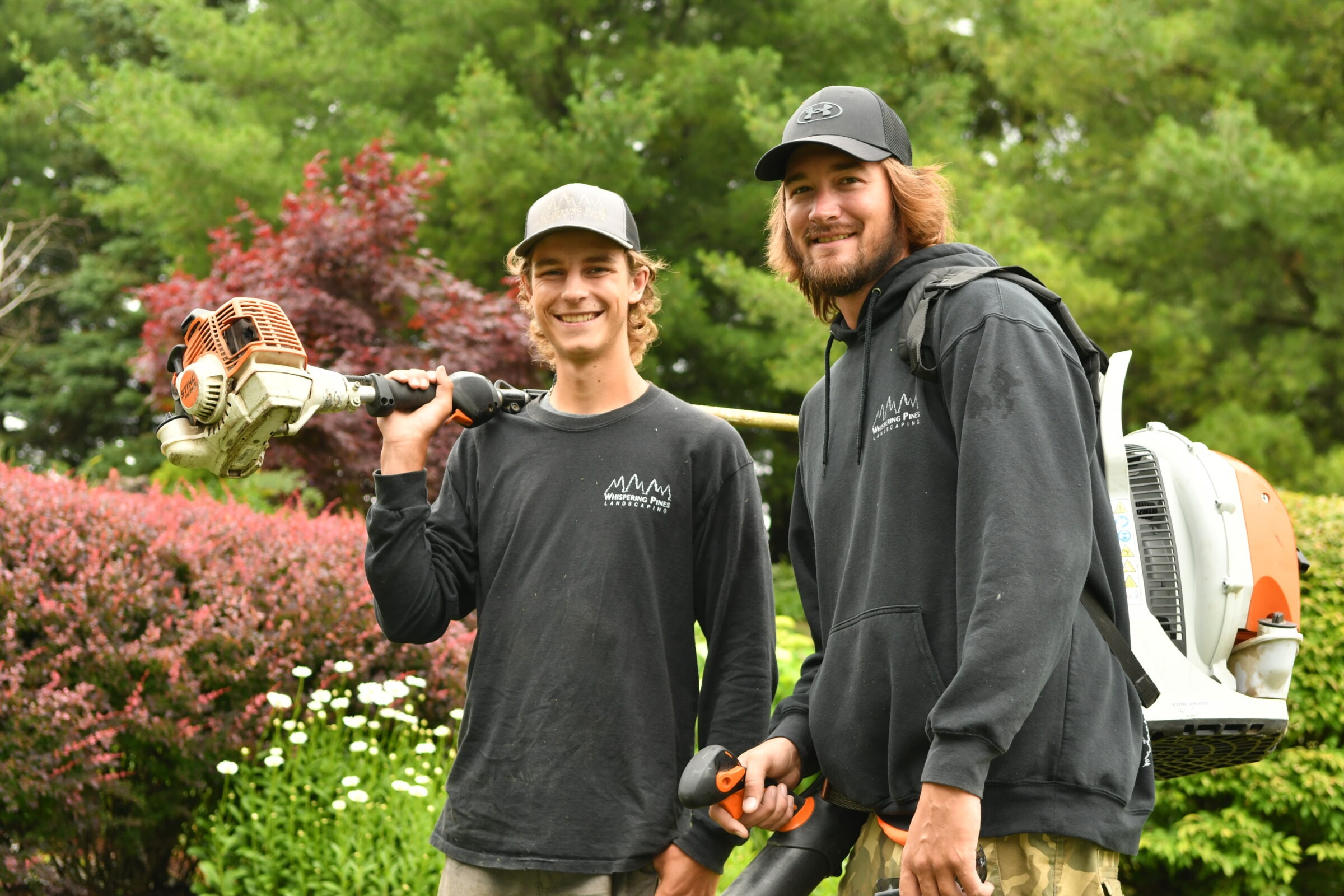 Two smiling persons in outdoor work attire, holding landscaping tools, are surrounded by vibrant greenery and flowers, conveying a cheerful, professional atmosphere.