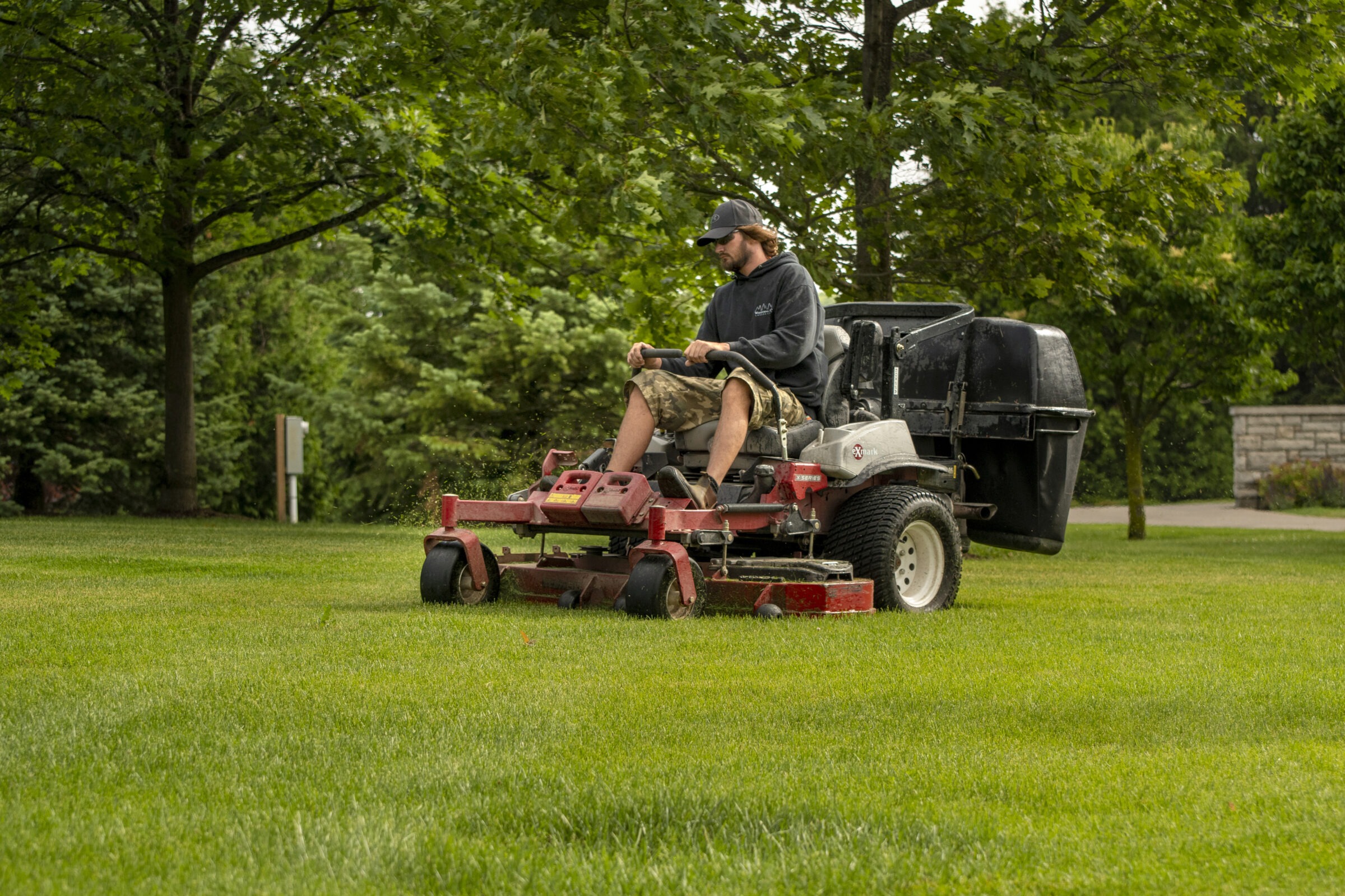 A person operates a red riding lawnmower, cutting grass in a lush, tree-filled park, wearing casual clothing.