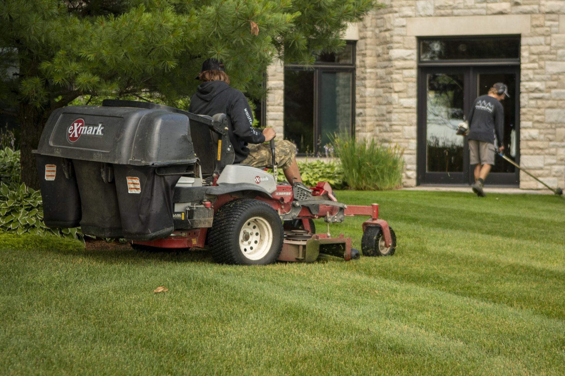 Two people maintaining a lawn; one using a riding mower, another trimming edges. They're working in front of a stone building.