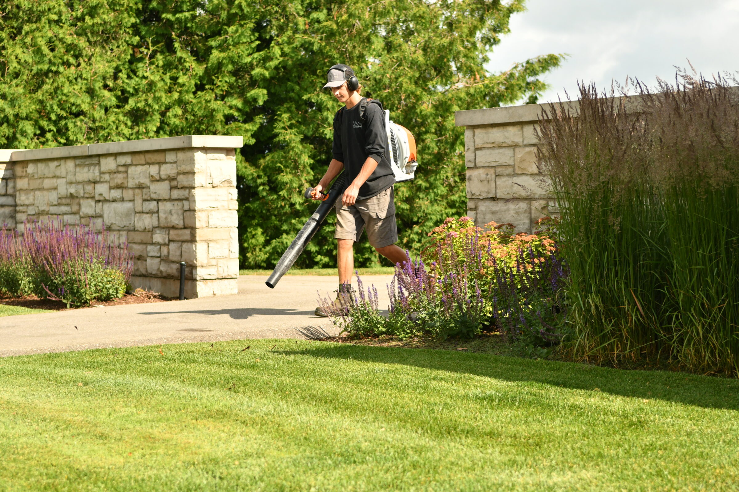A person with a leaf blower walks along a garden path, surrounded by greenery, stone walls, and colorful flowers on a sunny day.