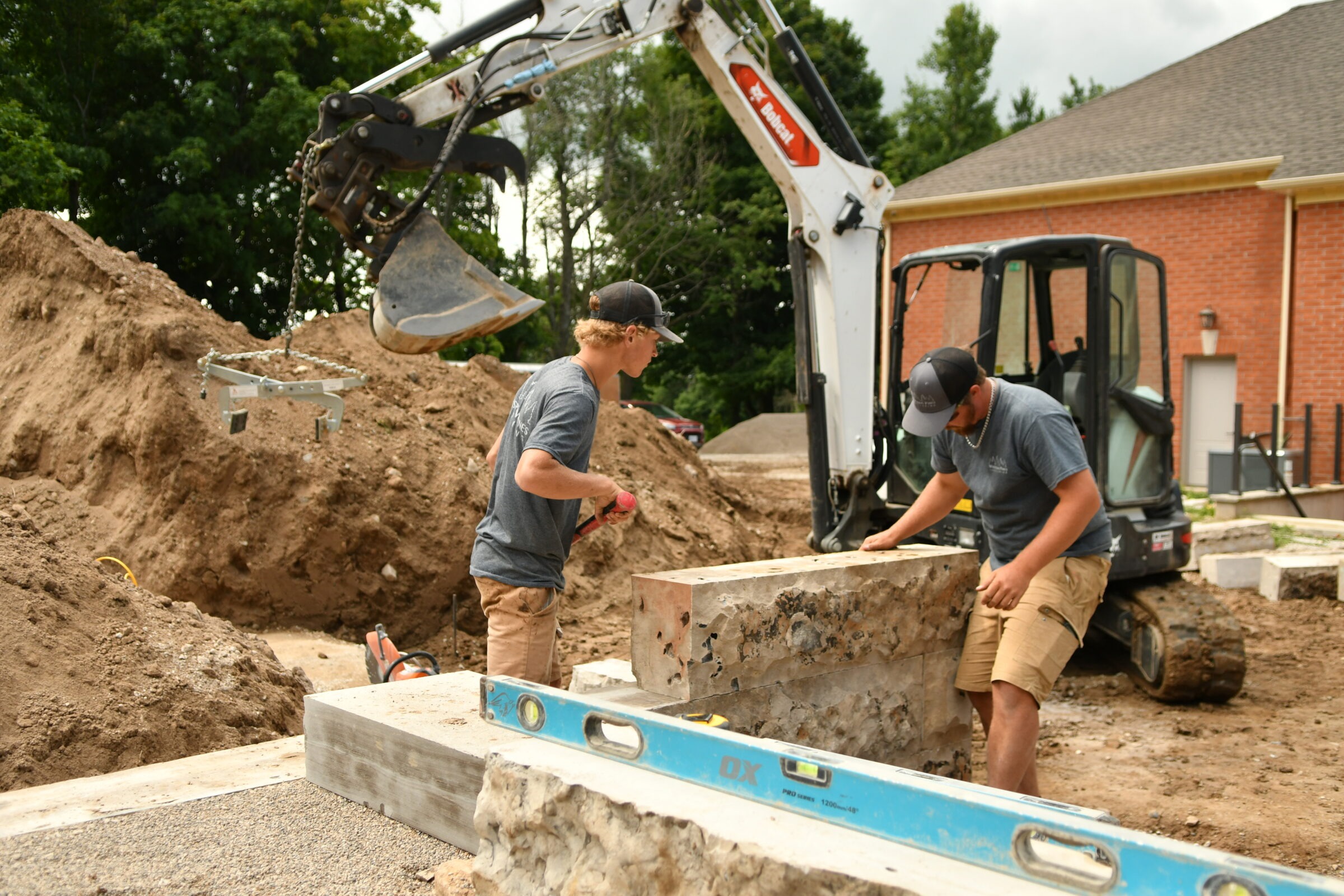 Two people work on a construction site, using heavy machinery and tools, surrounded by piles of dirt, beside a brick building.