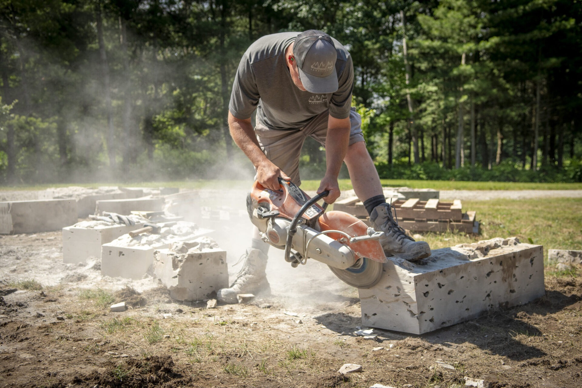 A person using a power saw cuts concrete outdoors, surrounded by dust and trees, on a sunny day with grass and stone blocks nearby.