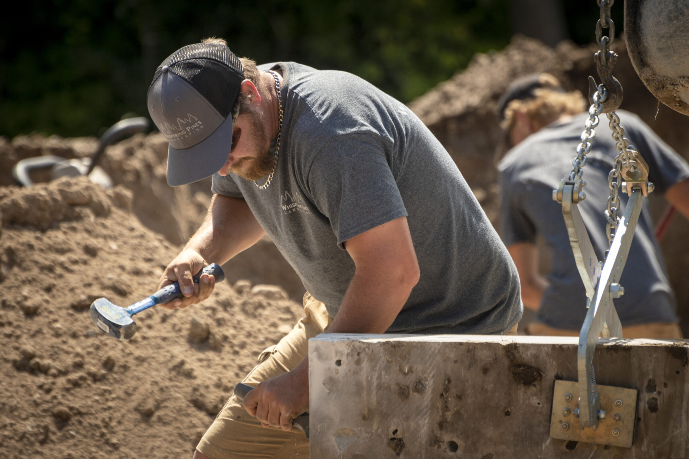 Two people working outdoors in a construction site, using tools on a large concrete block. Chains are visible for lifting.