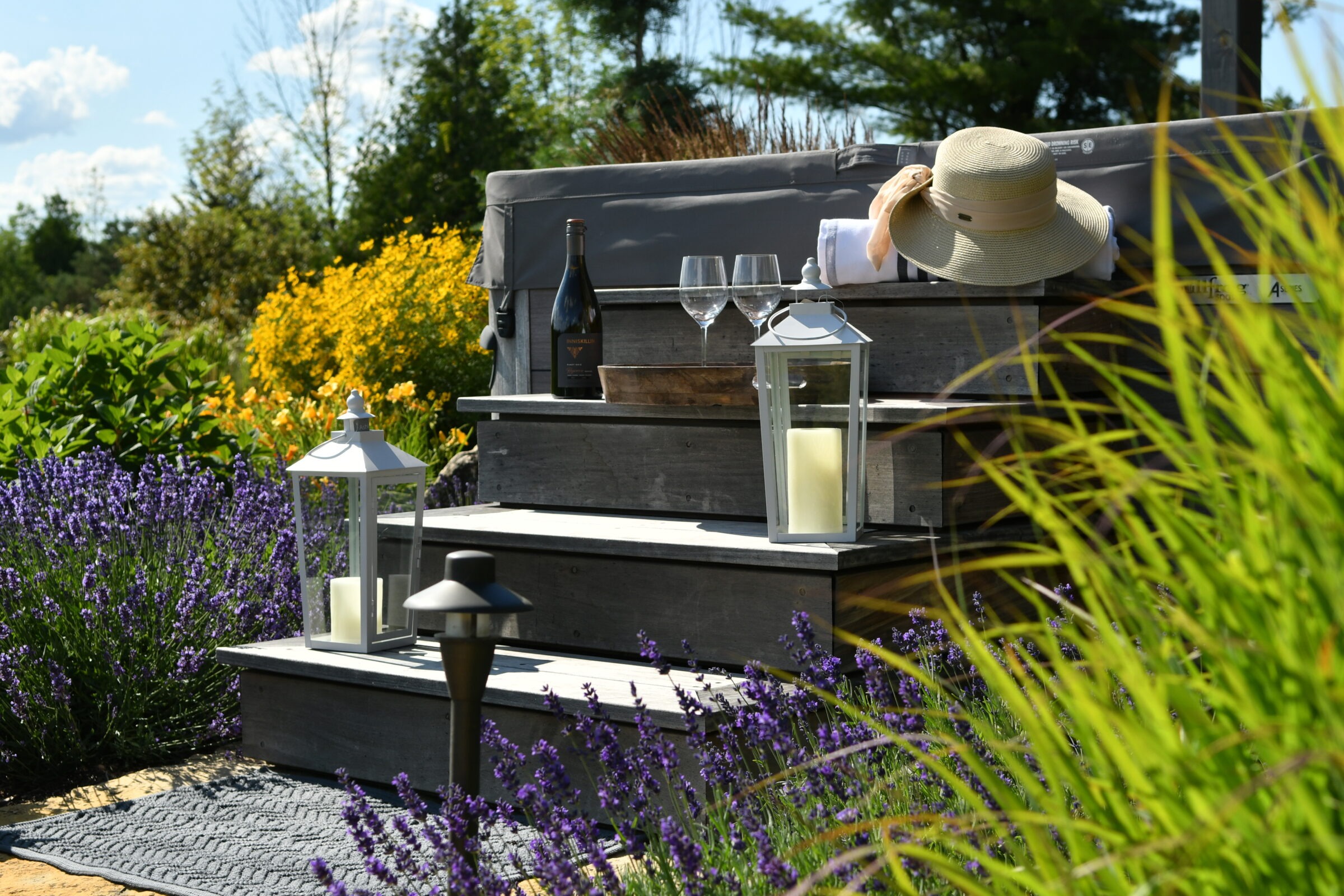 A serene garden setting with lavender, candles, a bottle of wine, glasses, and a hat resting on rustic wooden steps.