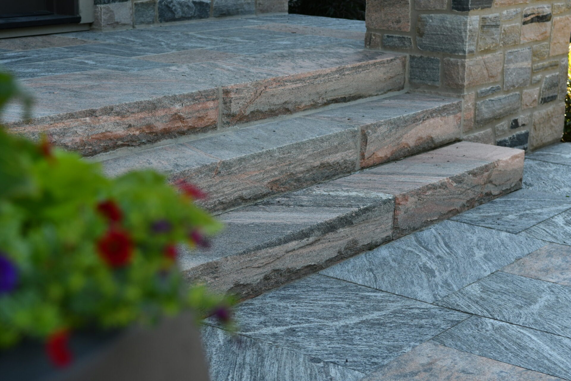 Stone steps and a textured wall are in focus, with blurred green plants and small red flowers in the foreground.