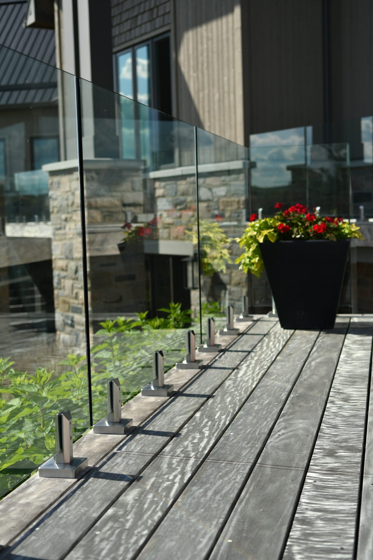 Modern terrace with glass railing, wooden floor, stone facade. Flower pots with red blooms add color. Peaceful outdoor space, no landmarks visible.