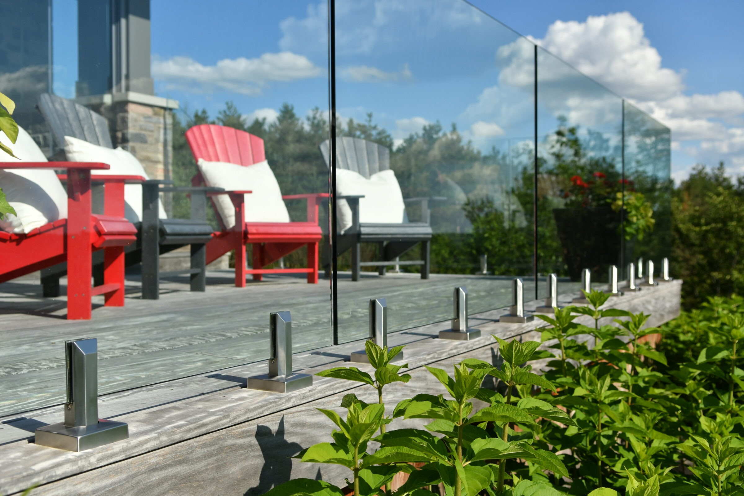 Modern terrace with red and gray chairs, glass railing, surrounded by lush greenery. Clear blue sky with scattered clouds overhead.