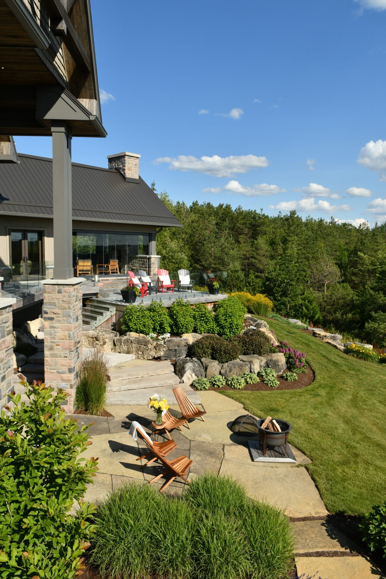 Modern house with a spacious patio, wooden furniture, fire pit, and vibrant garden, surrounded by lush greenery under a clear blue sky.