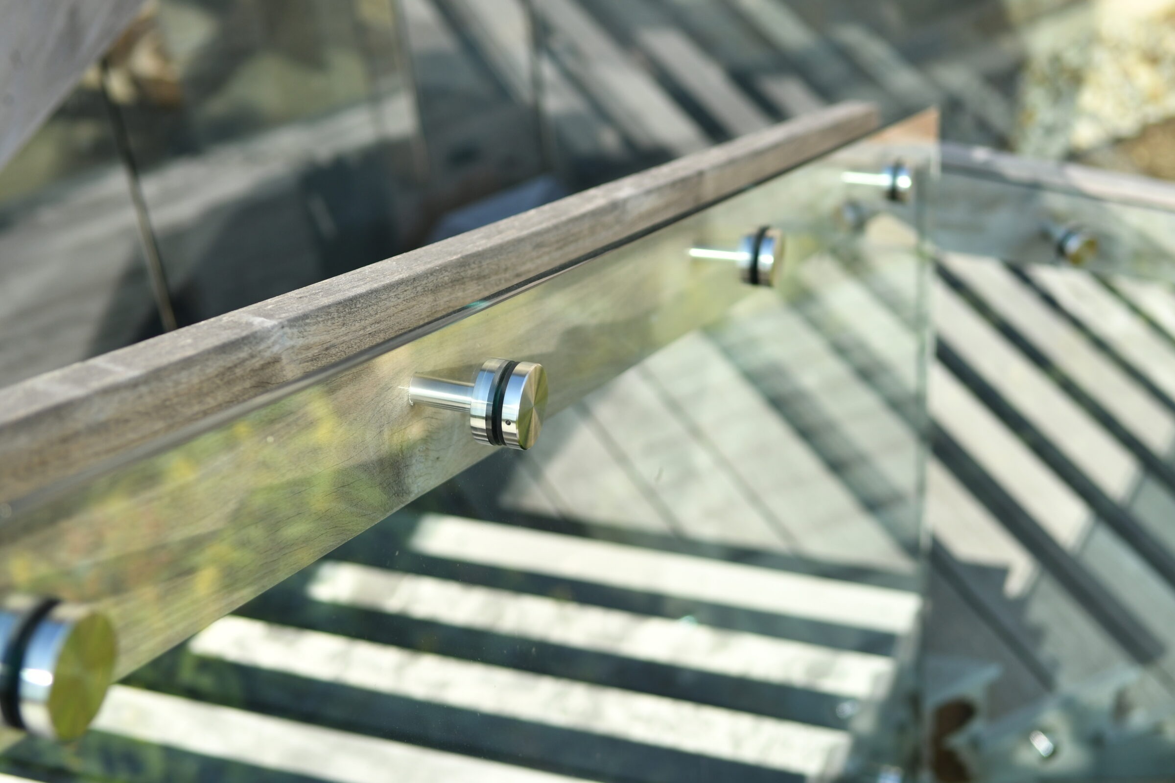 Close-up of a modern glass balustrade with metal fittings, casting striped shadows on a wooden deck in natural sunlight.