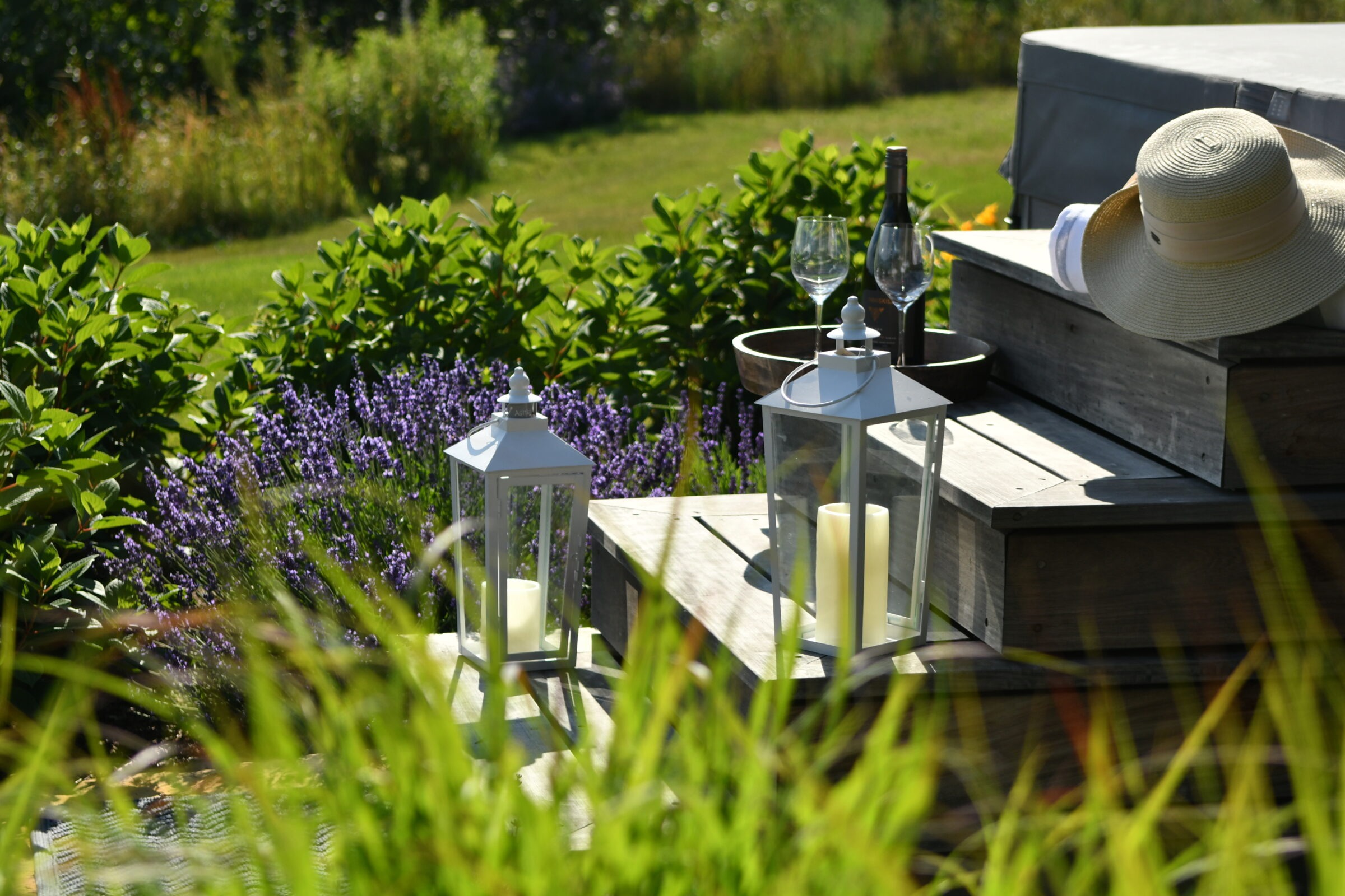 A sunlit garden with lavender, lanterns, wine glasses, and a hat on wooden steps. Peaceful outdoor setting with vibrant greenery.