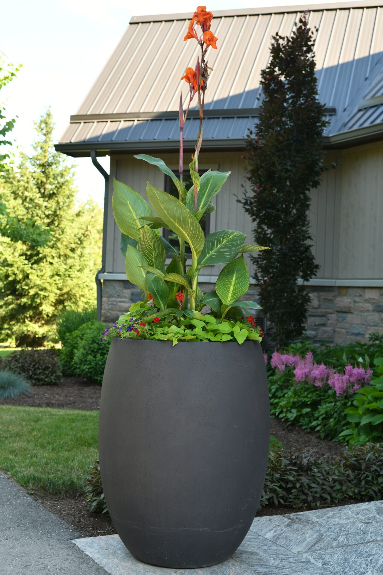 A large, dark planter with vibrant green plants and orange flowers in a garden setting, beside a modern building with metal roofing.