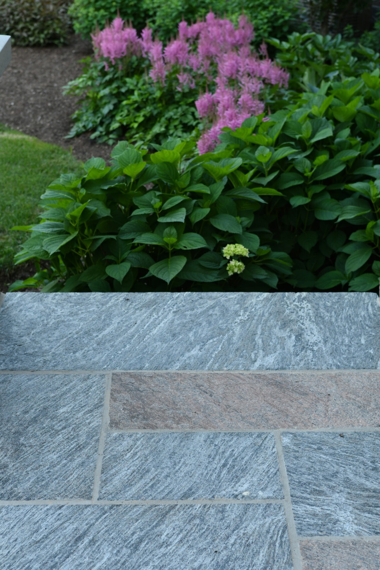 Stone patio with patterned tiles, bordered by lush green foliage and vibrant pink flowers, creating a serene garden setting.
