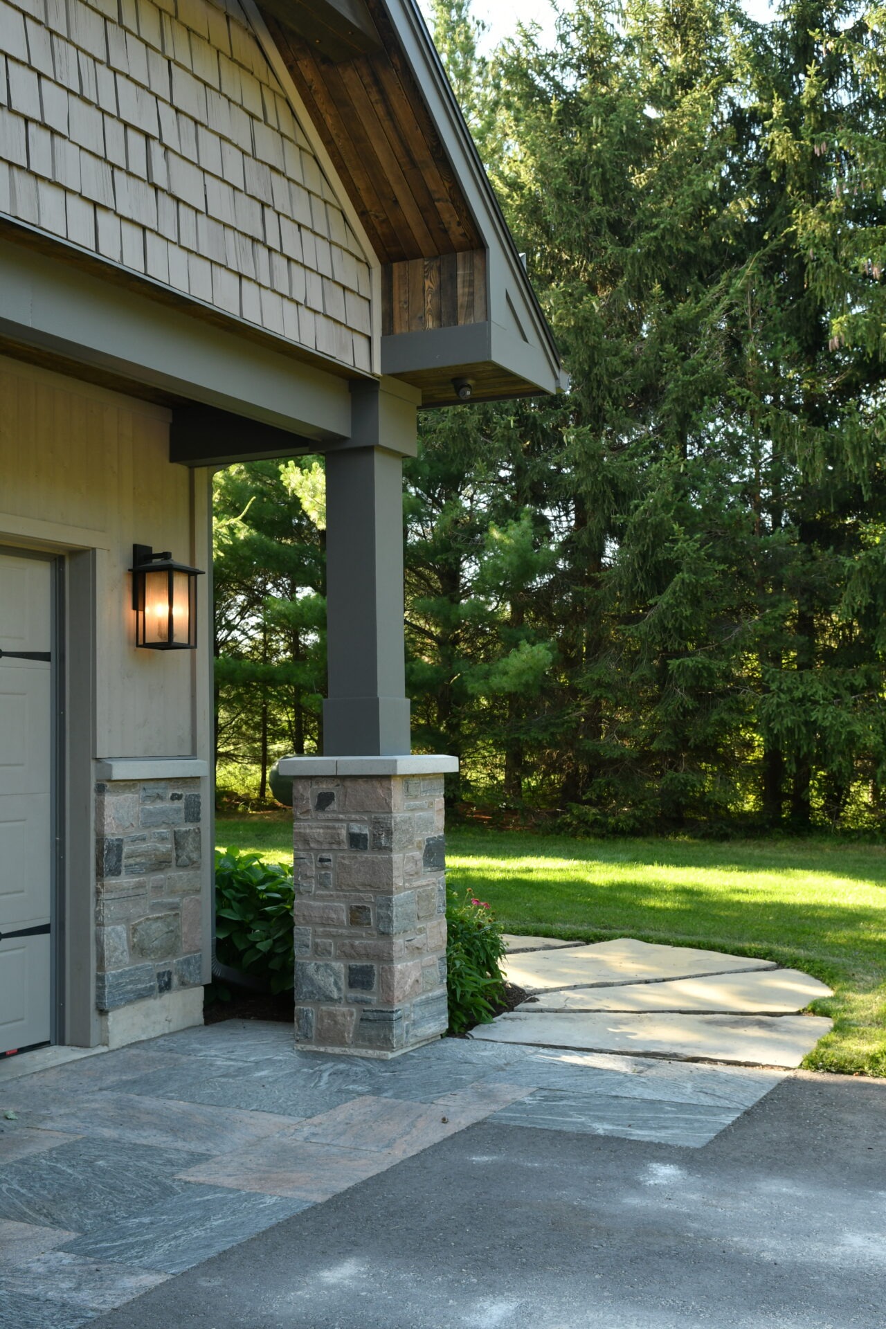A small patio beside a house with stone columns, lantern, and lush green lawn, surrounded by trees, creating a serene outdoor space.