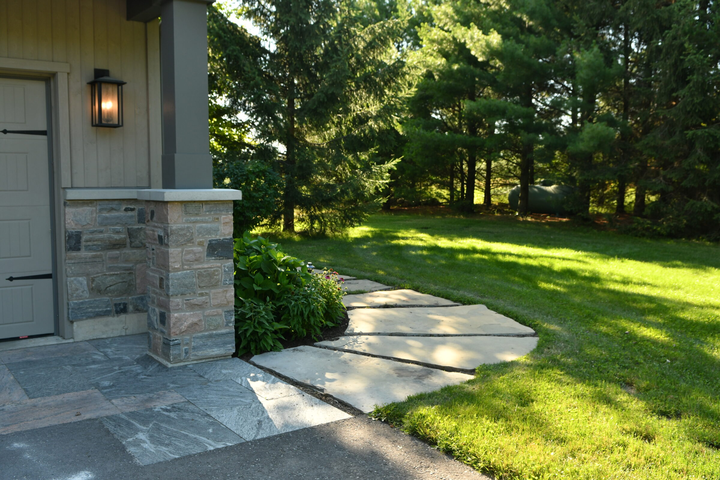 A sunlit garden path beside a stone house corner, surrounded by lush green grass and tall trees in the background.
