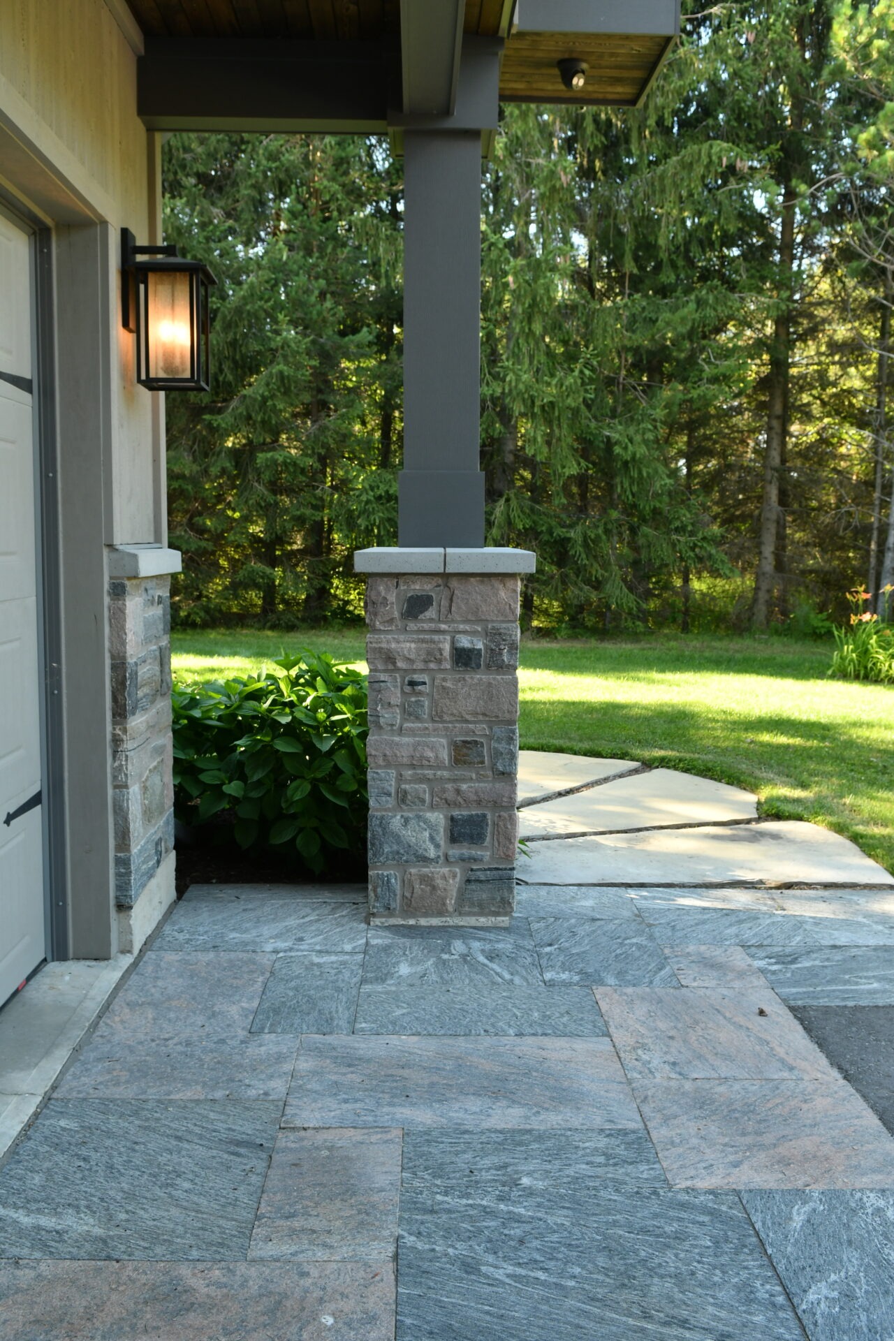 Stone patio with a lighted column under a wooden roof. Green shrubs and trees in the background create a serene, natural setting.