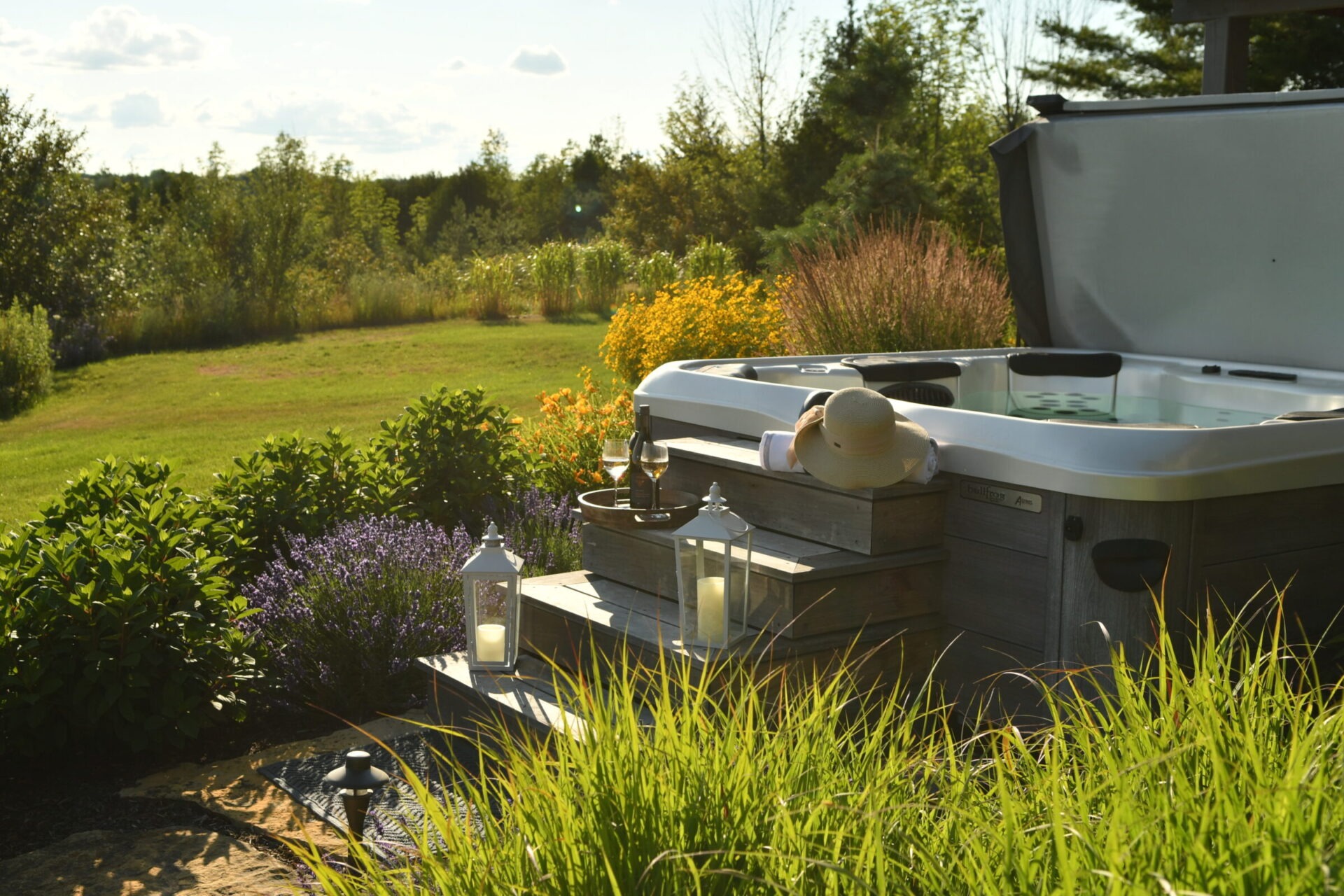 A person relaxes by an outdoor hot tub, surrounded by lush greenery and flowers, with wine glasses and lanterns nearby.