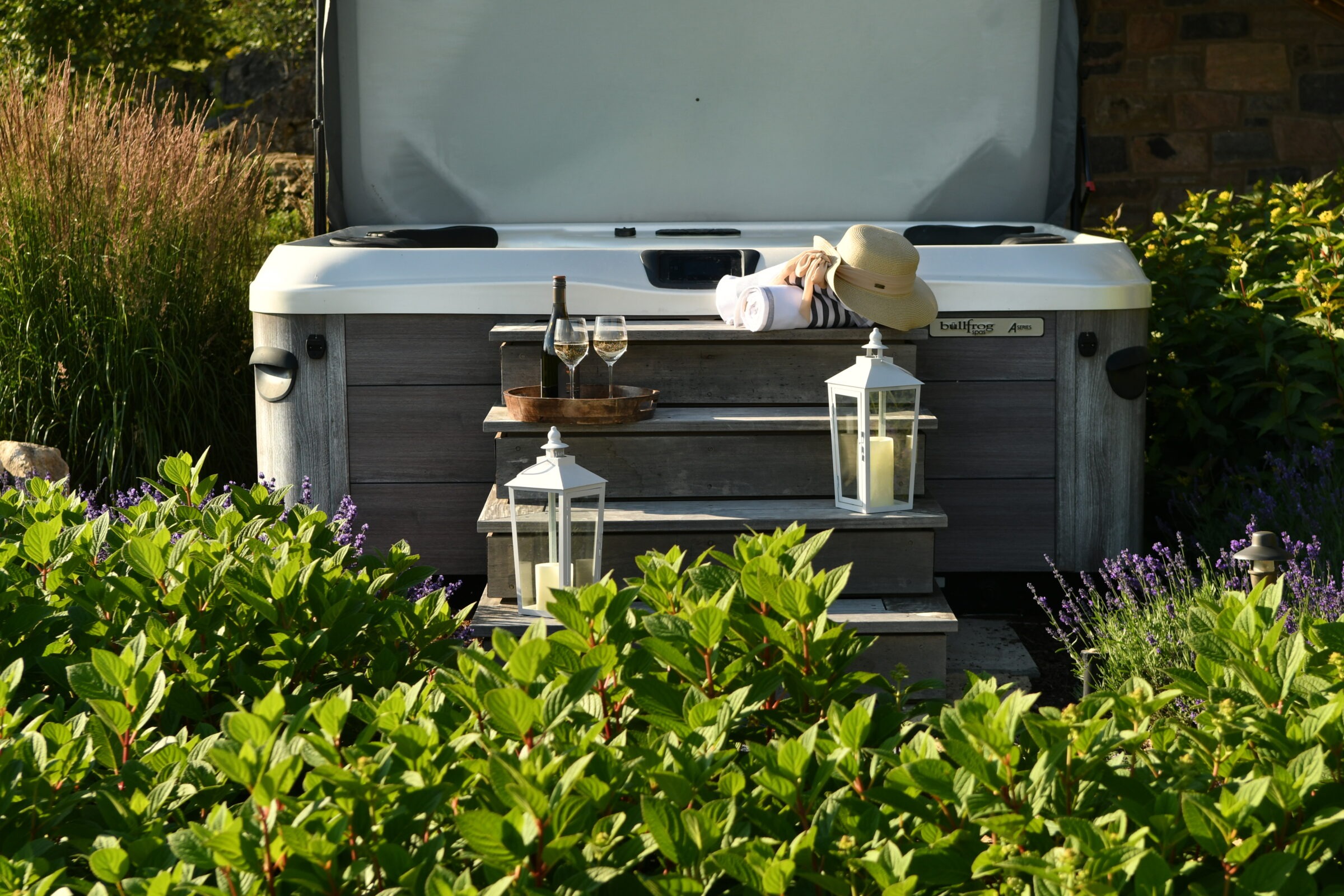 A person in a hat rests on a hot tub step surrounded by foliage, candle lanterns, wine bottle, and glasses; peaceful outdoor setting.