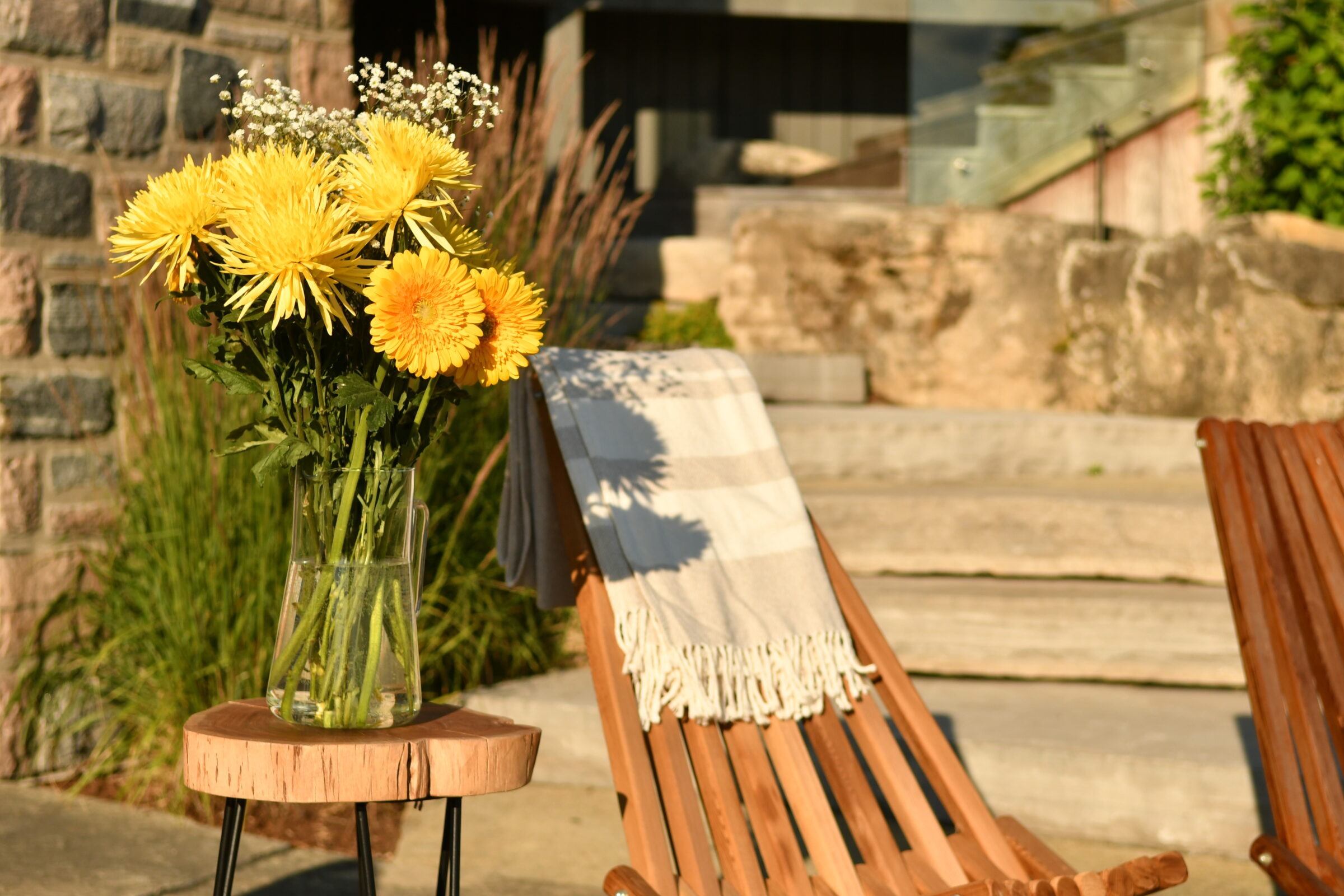 Yellow flowers in a glass vase sit on a wooden table beside a deck chair with a blanket, outdoors in sunlight.