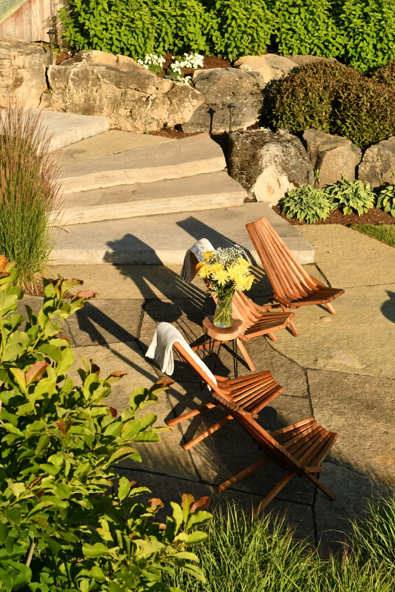Sunlit garden patio with wooden chairs and a table, featuring a vase of yellow flowers. Surrounded by rocks, greenery, and stone steps.