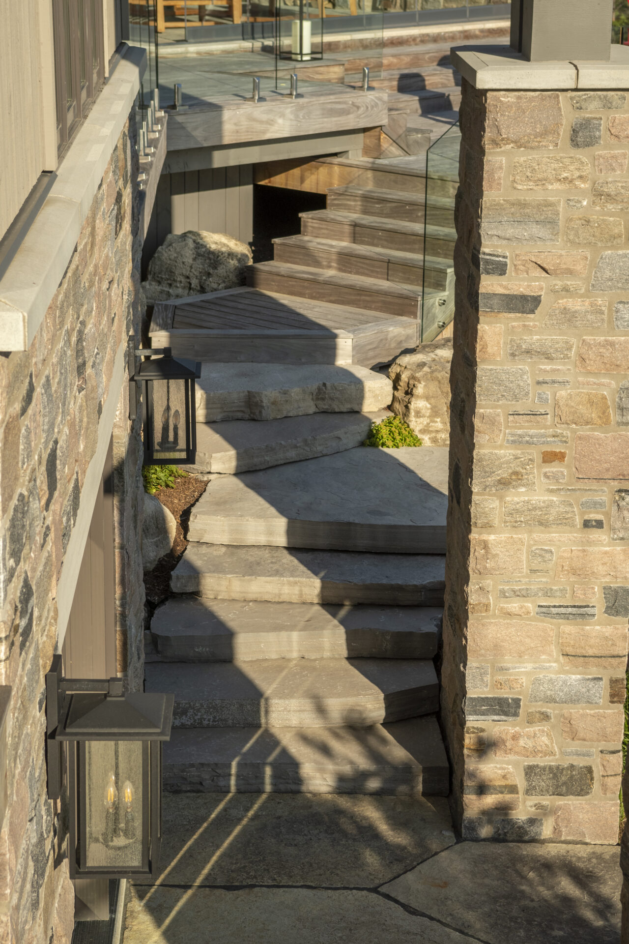 A rustic, sunlit stone staircase with modern, glass railing, flanked by walls, leading to a small patio area.