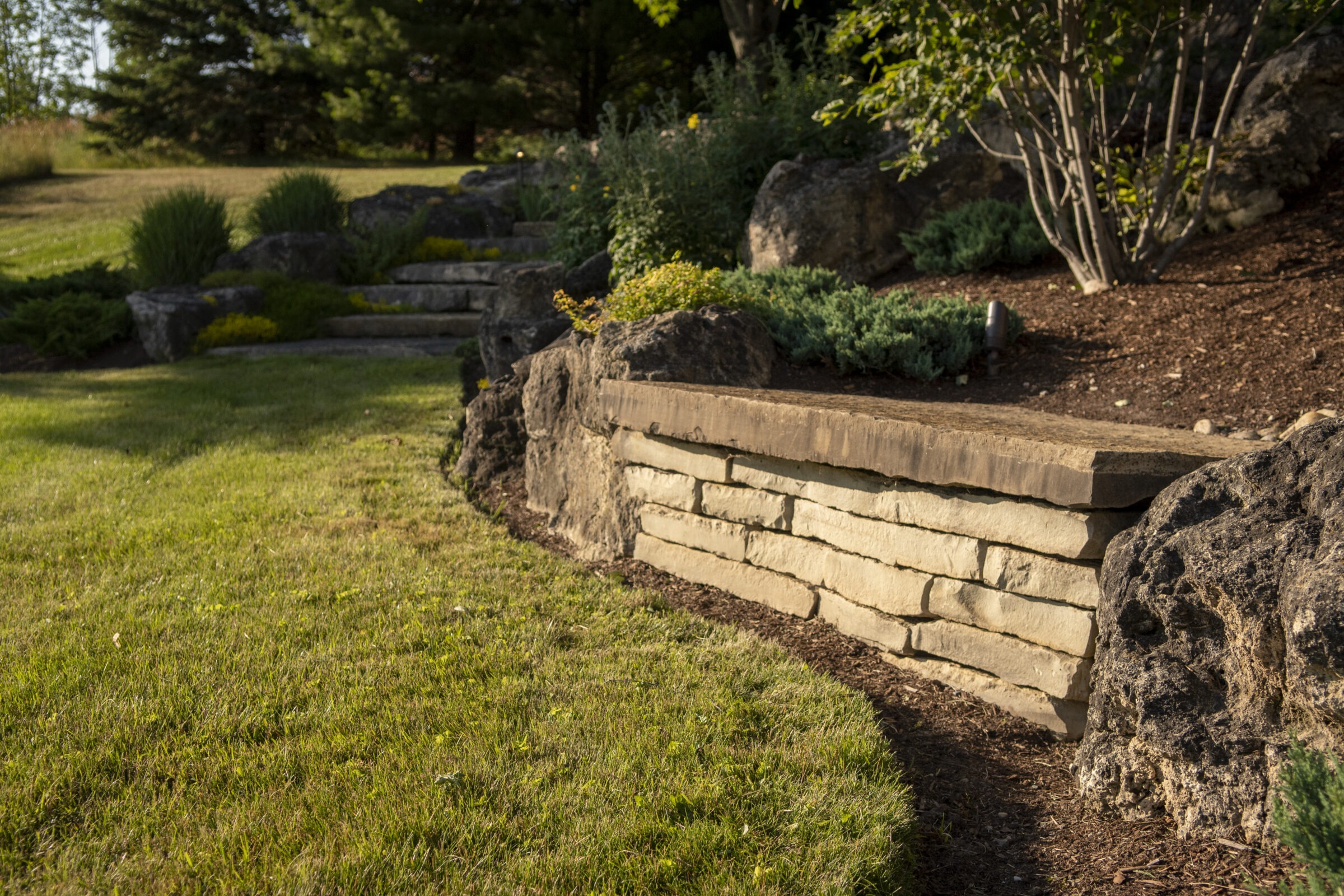 A serene landscape featuring a stone retaining wall, lush greenery, and trimmed grass under soft, natural sunlight, with shadows indicating early morning or evening.
