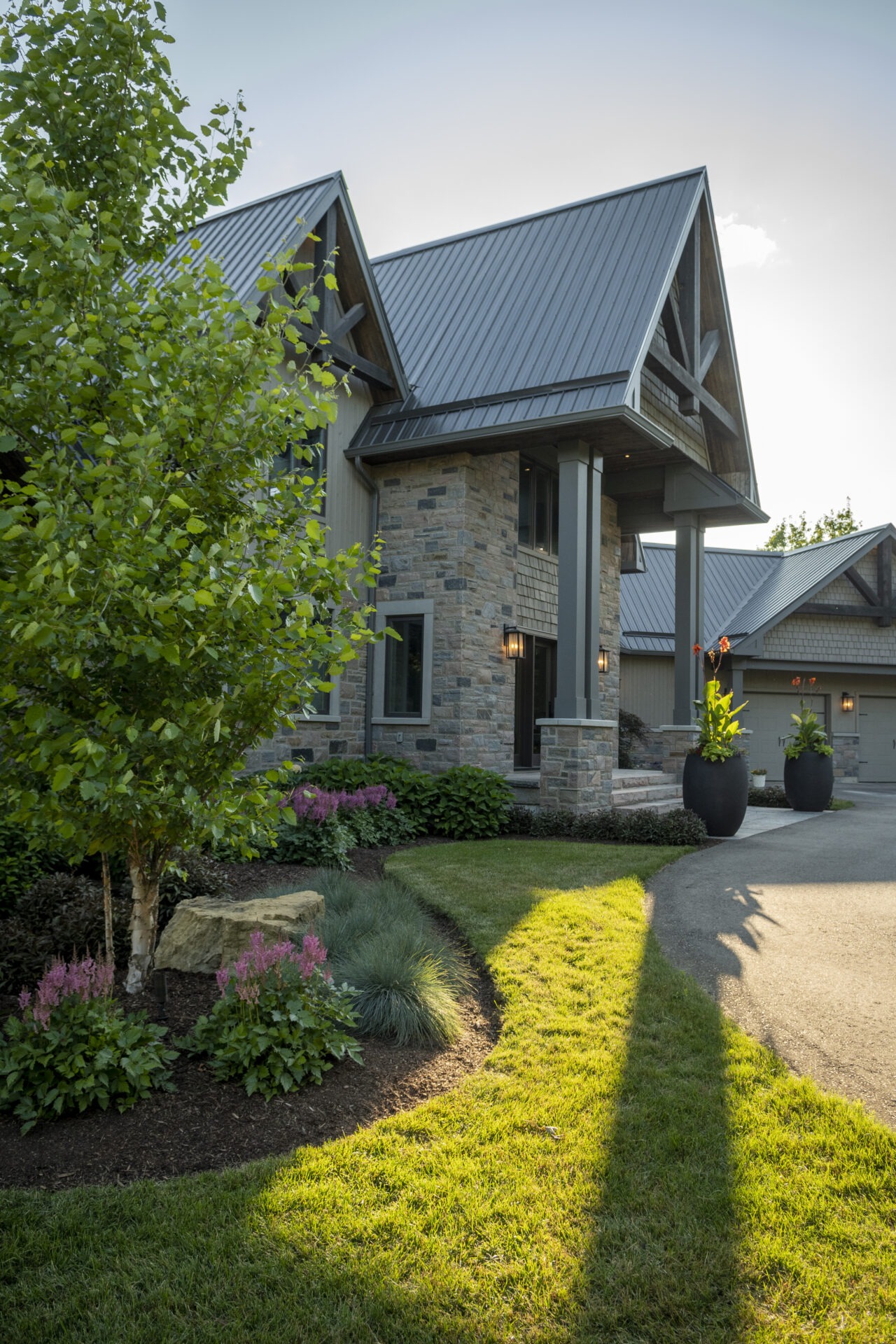 Modern stone house with gabled roof, lush landscaping, and sunlit pathway. Two black planters with tall plants flank the entrance.