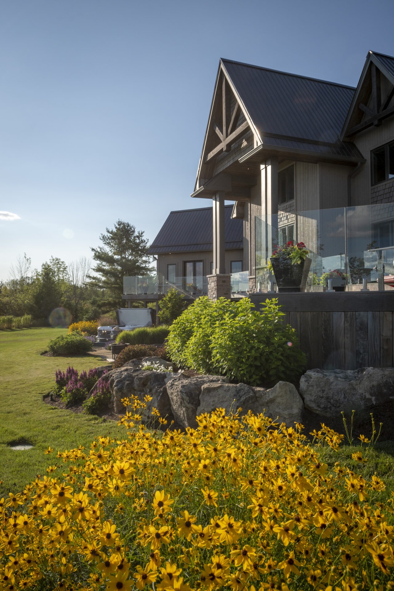 A modern, stone-house with a glass balcony overlooks a vibrant garden with yellow flowers on a sunny day.