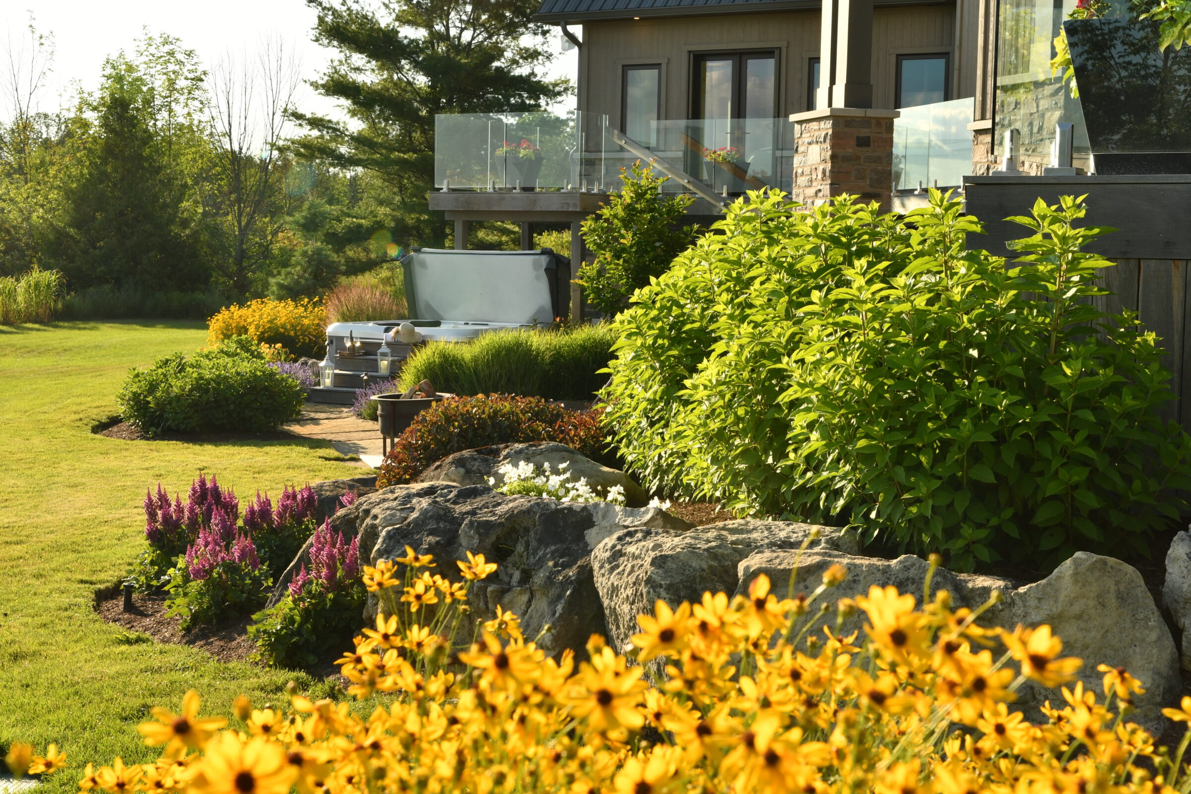 A garden with vibrant flowers, lush greenery, and a modern patio beside a house with glass railings under the bright sunlight.