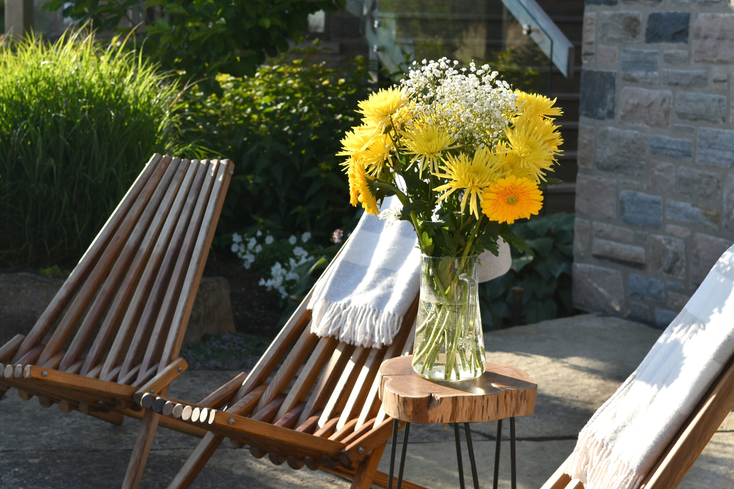 A vase of vibrant yellow flowers on a small table flanked by wooden chairs, set outside amid lush greenery and rustic stone walls.
