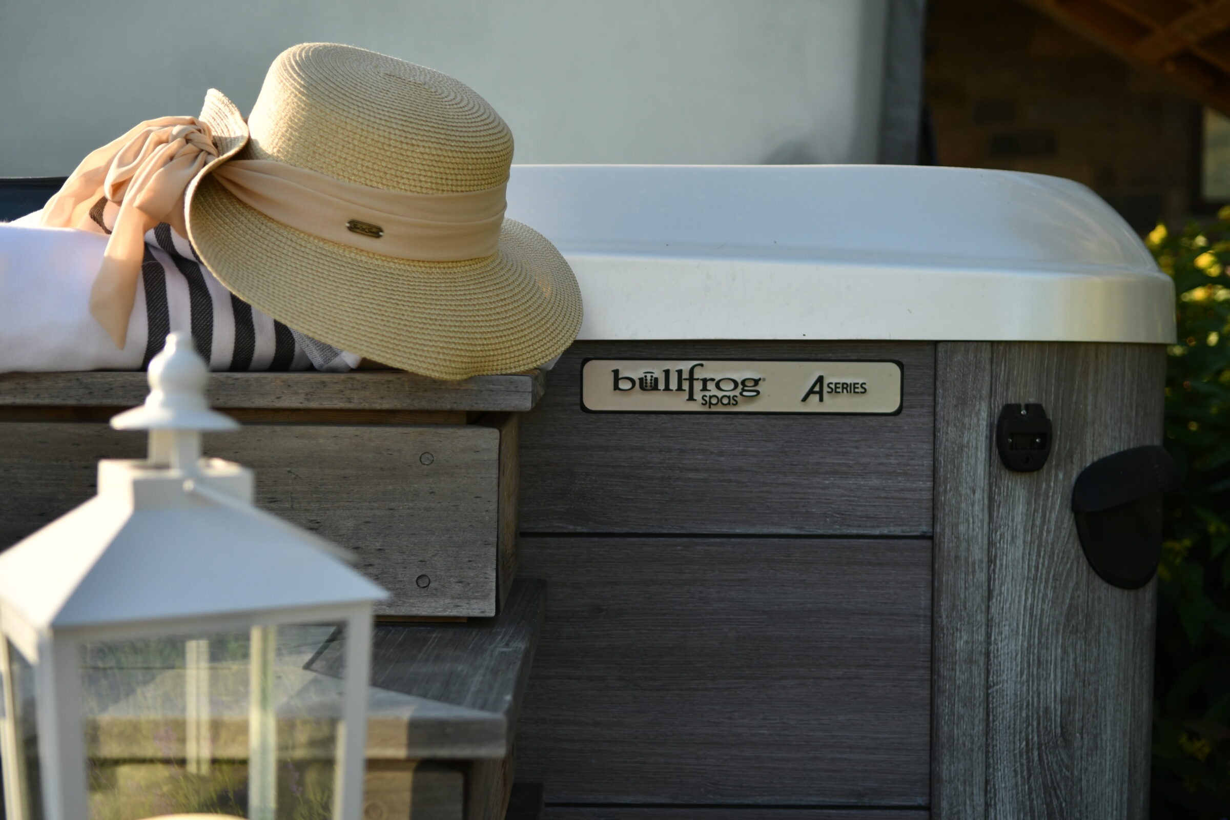 A straw hat and towel rest on wooden steps beside a Bullfrog Spas hot tub, with a white lantern in the foreground.