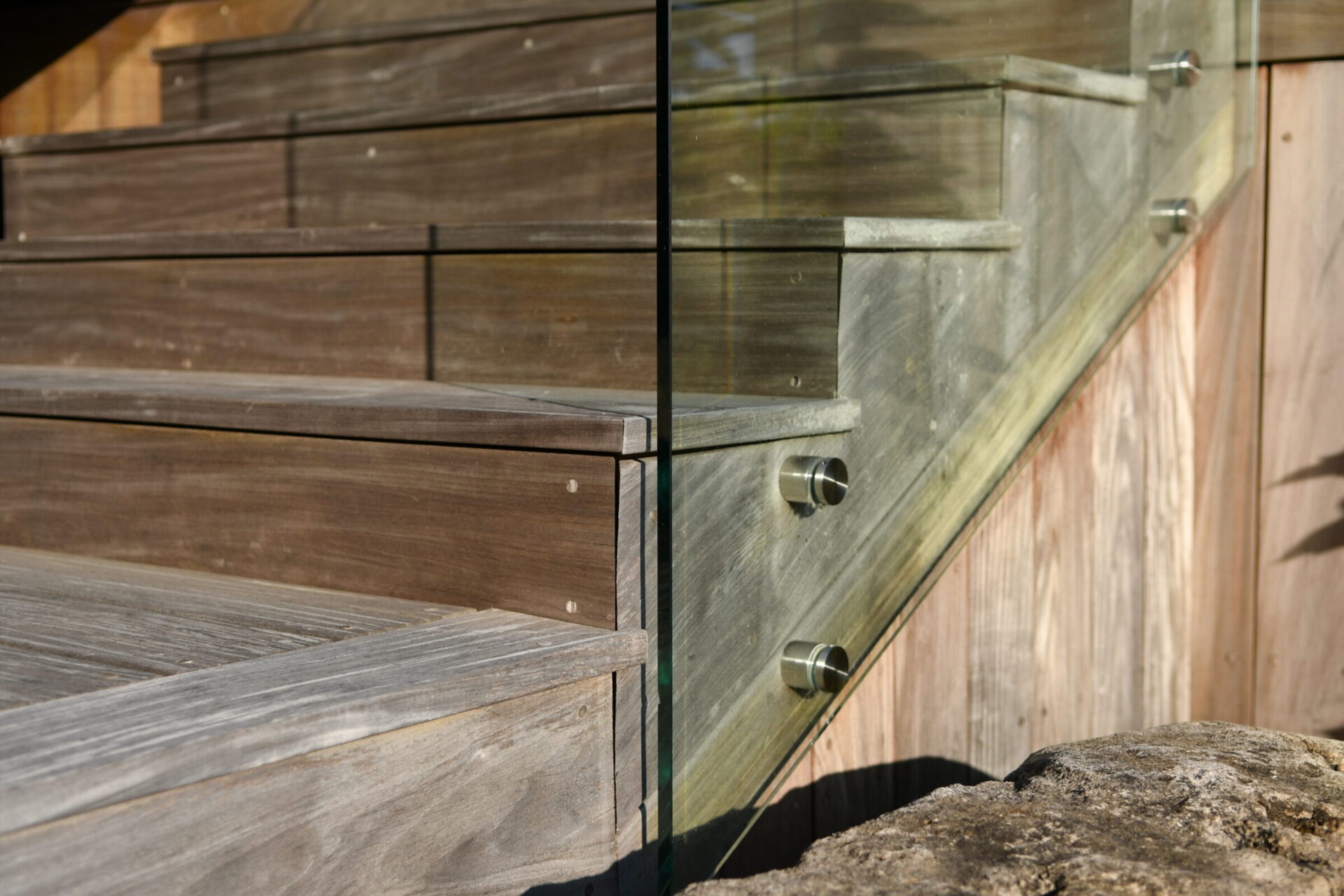 Wooden staircase with glass railing detail, reflecting sunlight. Stone elements at the base add natural contrast and texture to the modern design.