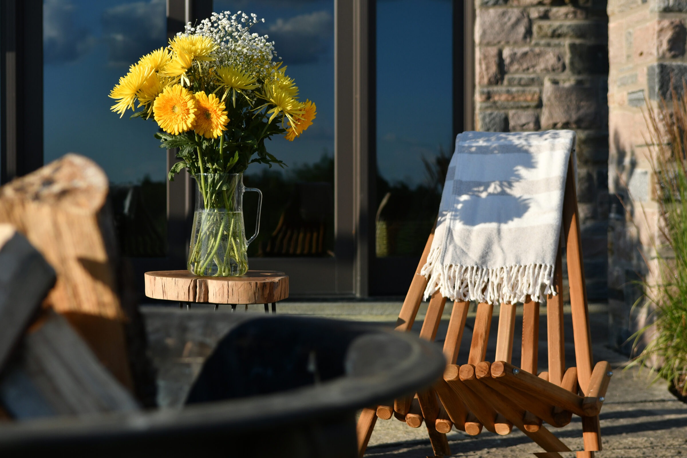 A sunny porch scene with yellow flowers in a glass jug on a small table, next to a wooden chair draped with a blanket.