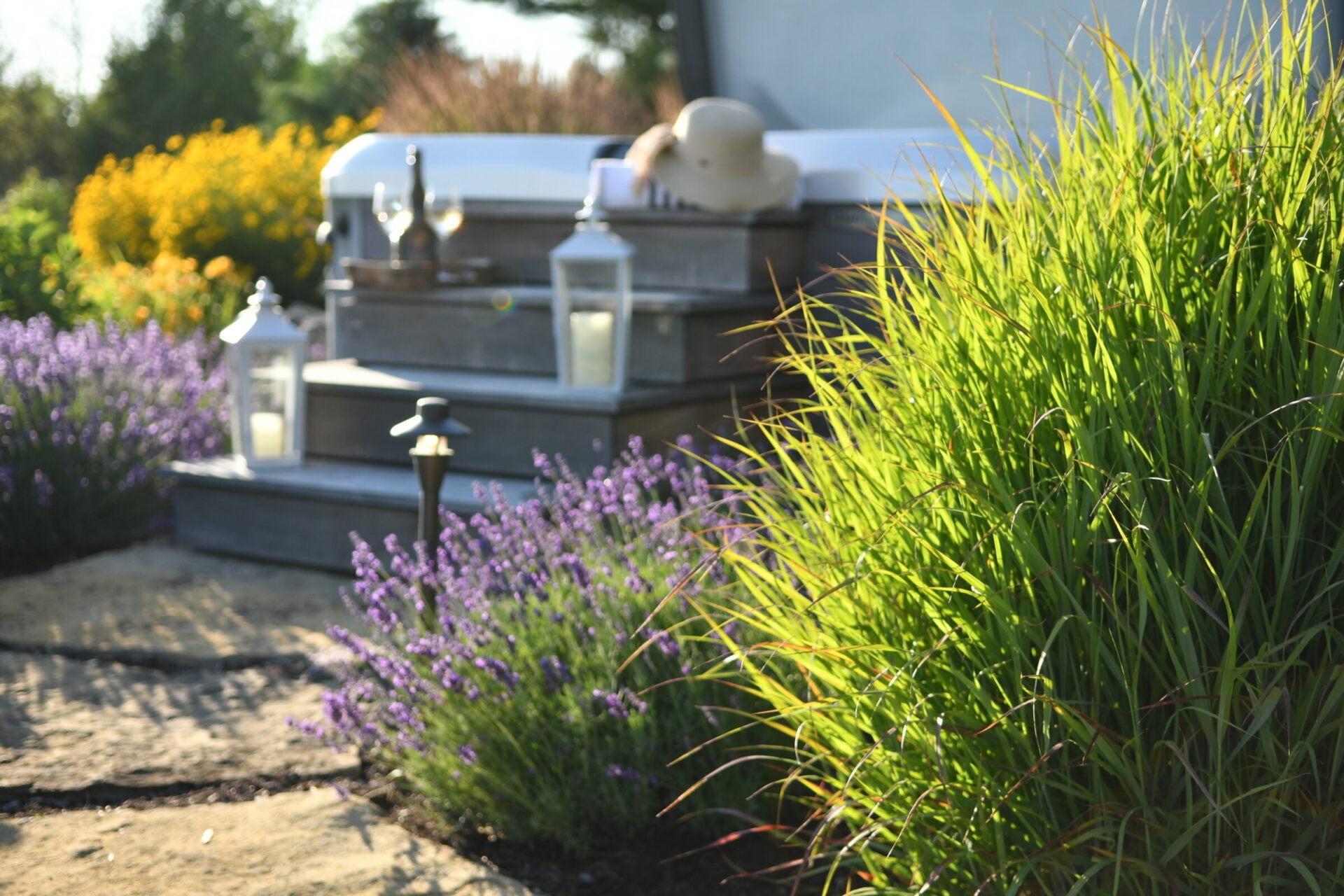 Garden with lavender, lush grass, hot tub with hat, two lanterns, and drinks on steps, under bright sunlight.