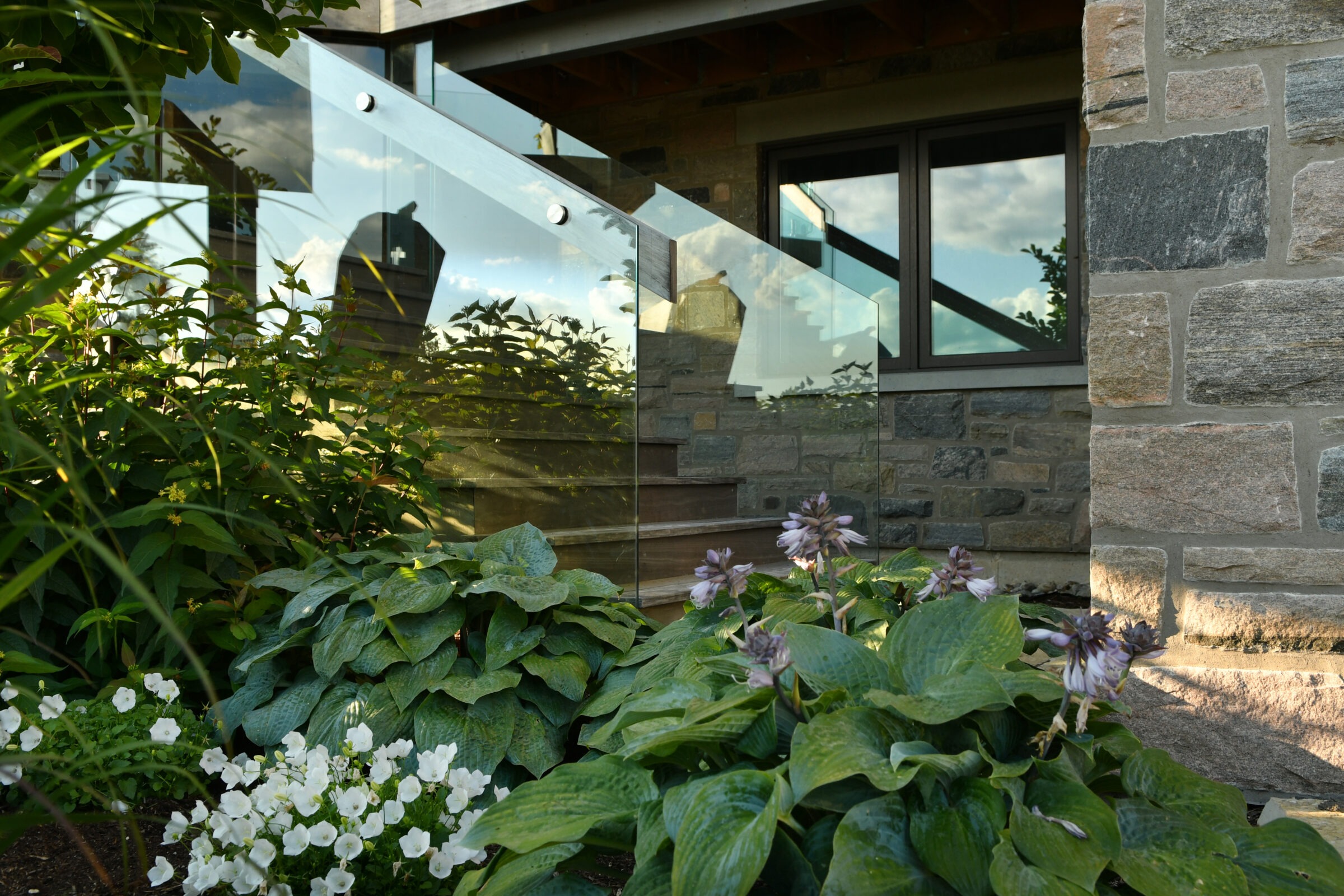 Stone building with glass railing reflects nature. Lush garden with hostas and white flowers enhances the peaceful, modern architectural design.