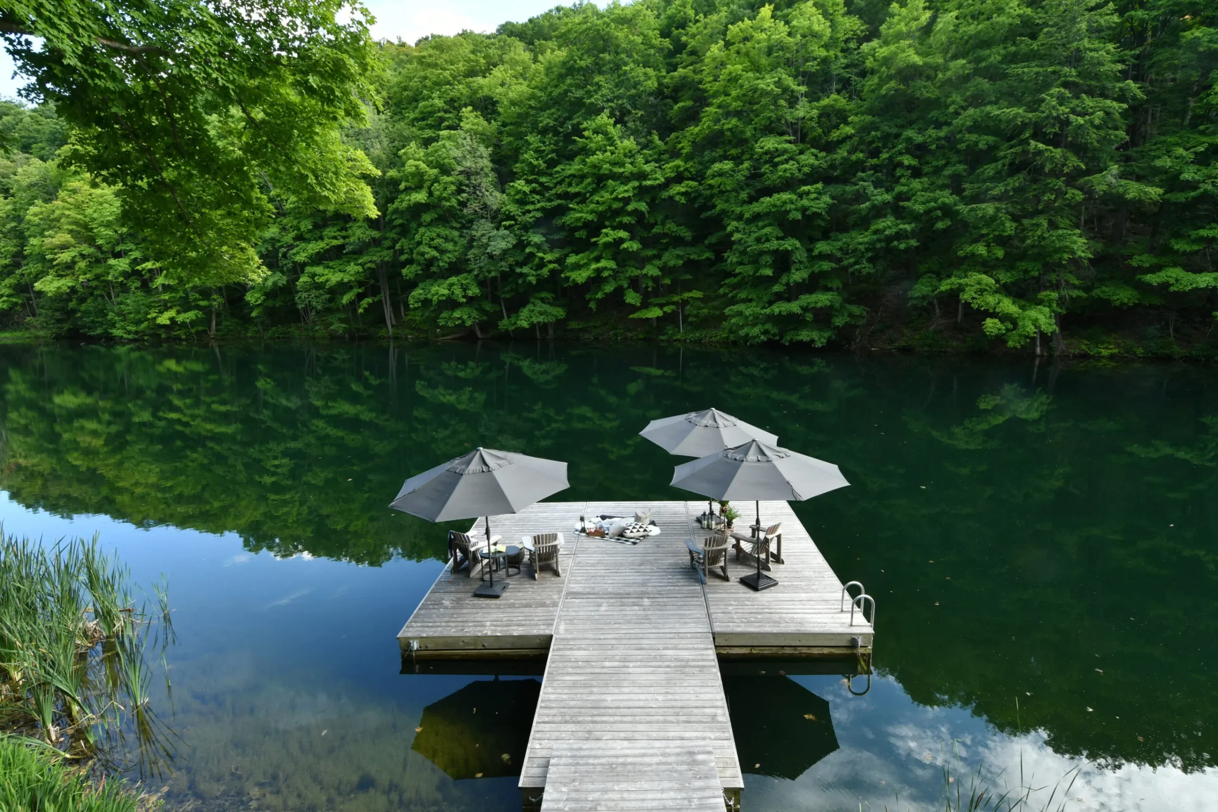 A tranquil wooden dock with umbrellas overlooks a calm lake surrounded by lush green forest reflections, creating a serene and peaceful atmosphere.