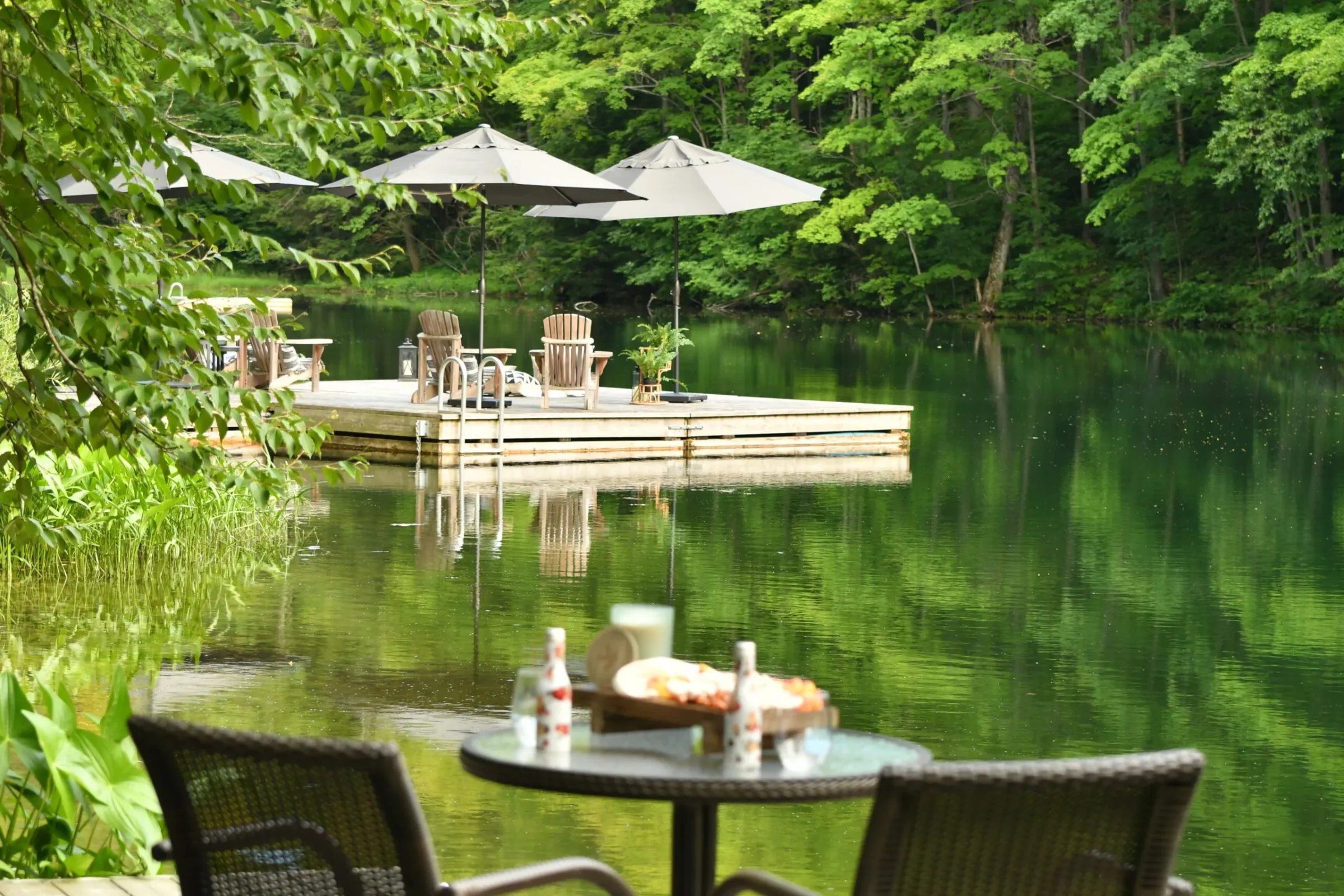 A serene lakeside scene with a wooden deck, loungers, and umbrellas. Foreground features a table with food and drinks. Lush greenery surrounds.