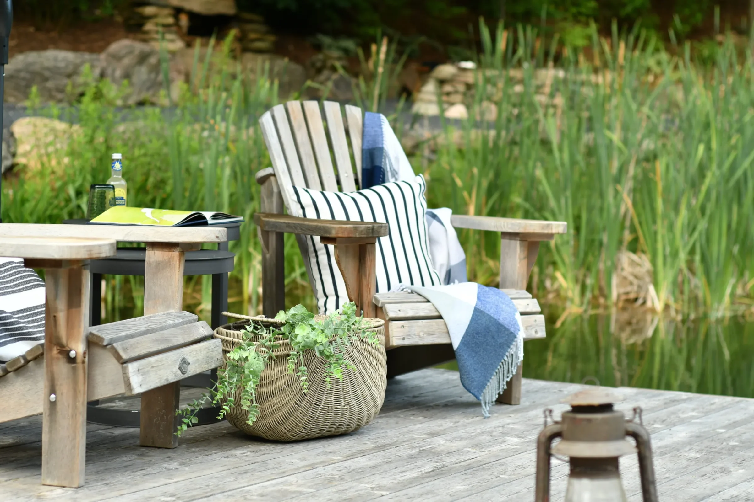 A wooden chair with cushions and a blanket on a lakeside deck, surrounded by lush greenery and a serene atmosphere.