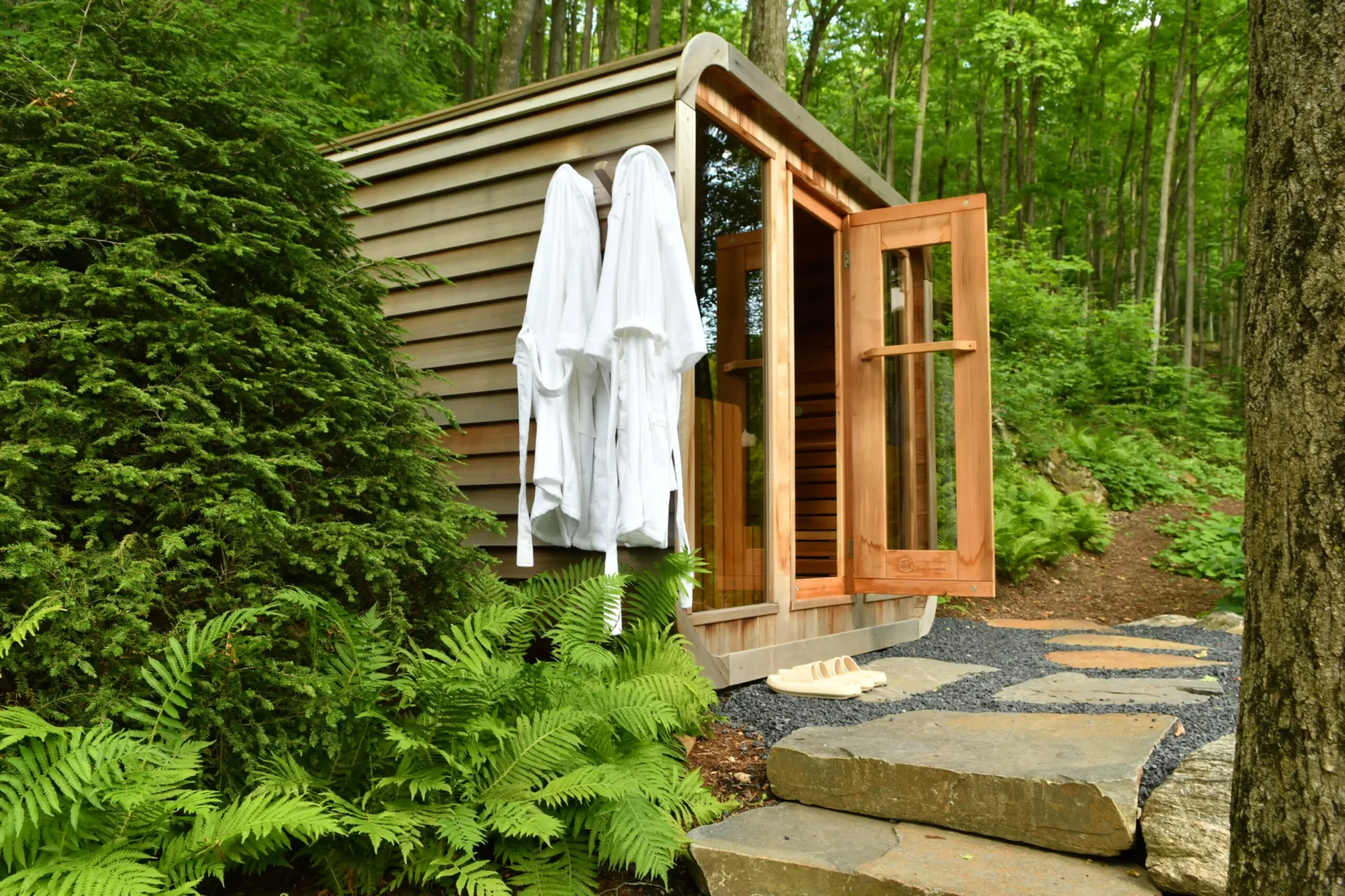 A wooden sauna with open doors, surrounded by lush greenery in a forest setting. Two white robes hang outside.