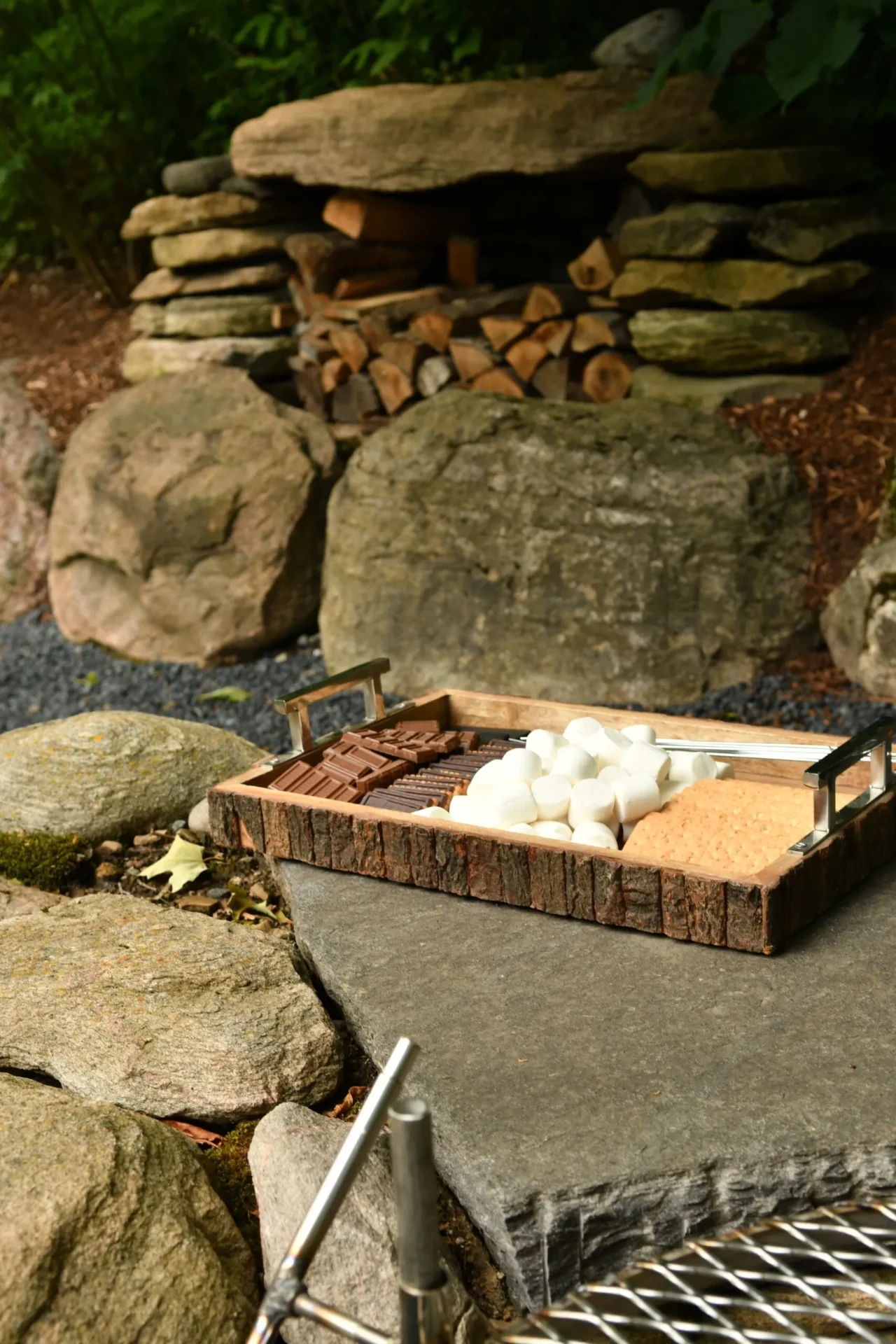 A tray with marshmallows, chocolate, and graham crackers sits on a stone near a neatly stacked wood pile in a forest area.