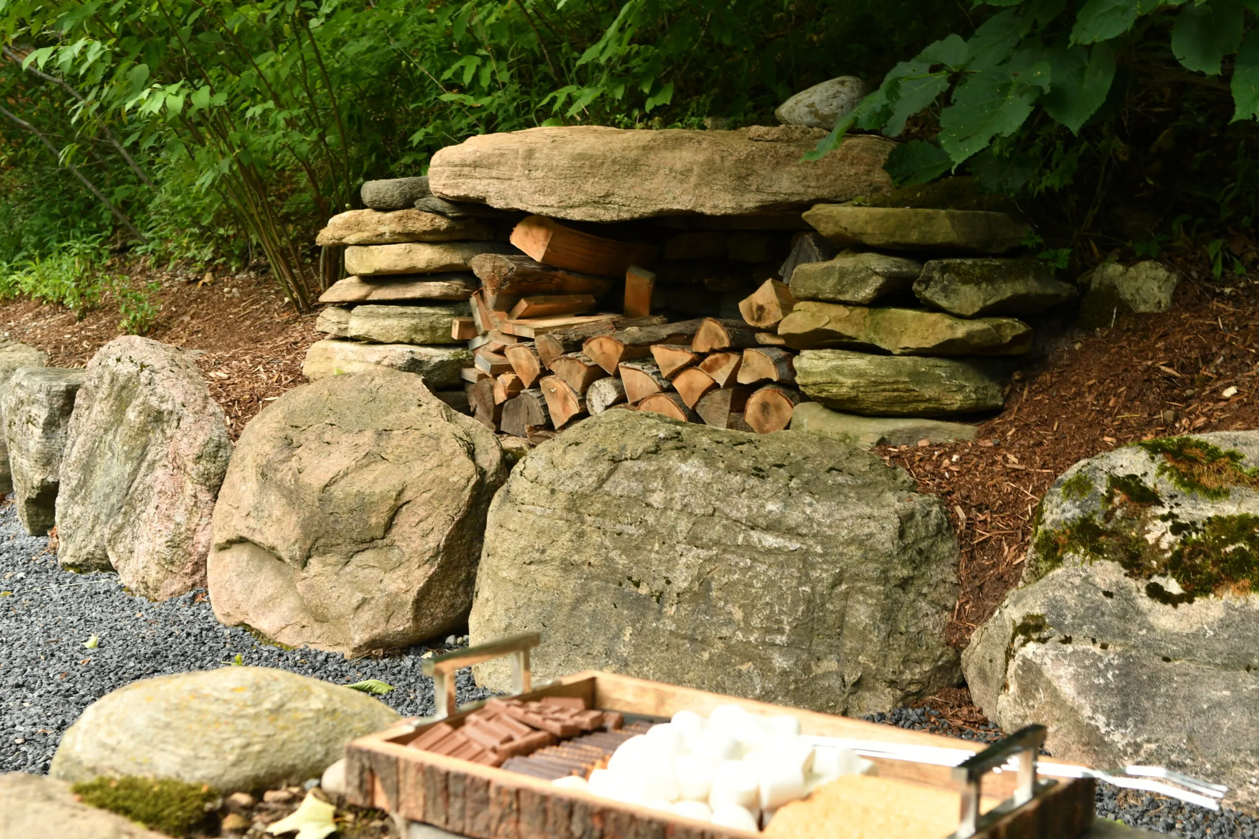 A natural stone woodpile holder surrounded by rocks, with a tray of chocolate, marshmallows, and graham crackers in the foreground.