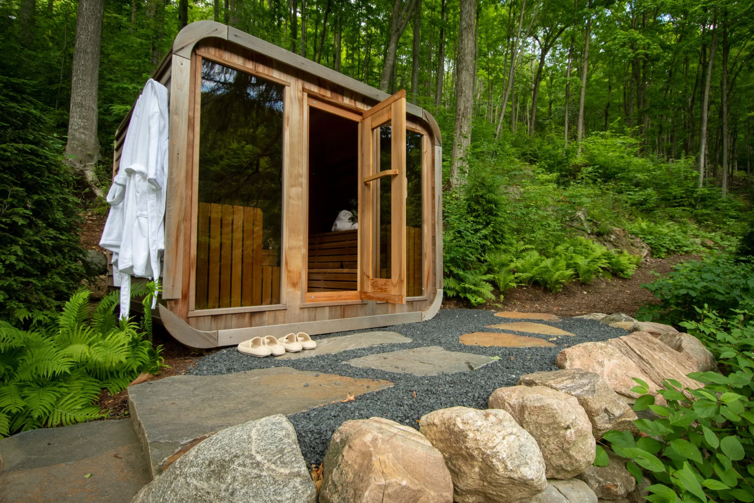 A wooden sauna with open door in a forest setting, path of stones and gravel leading to it, surrounded by lush greenery.