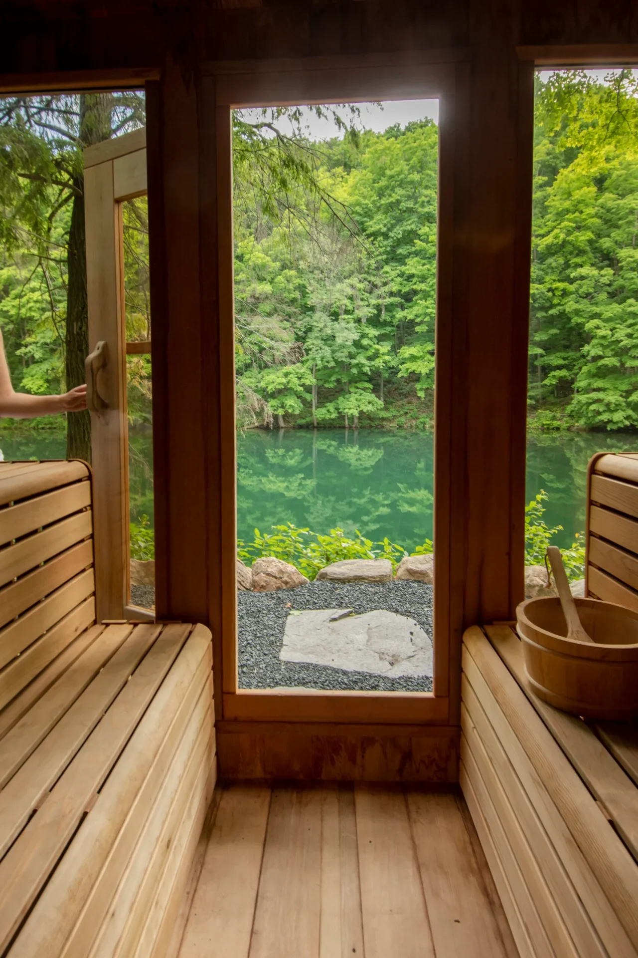 Wooden sauna with benches and bucket, open door showing lush green forest and tranquil blue lake outside. A person’s hand holds the door.