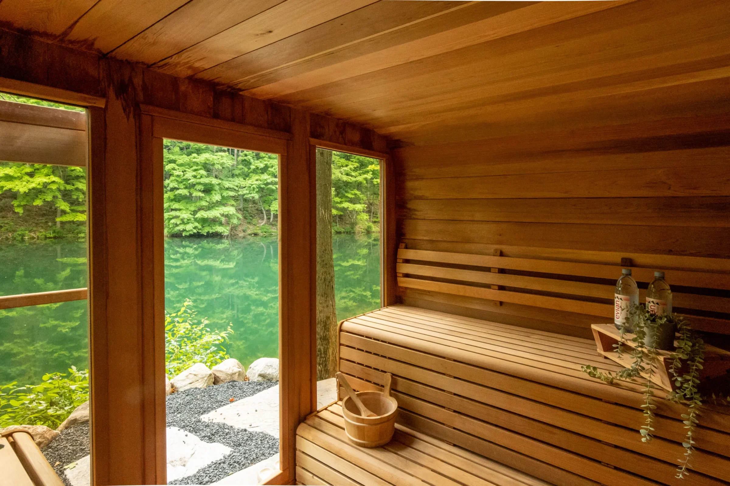 Wooden sauna interior with benches and a bucket, overlooking a serene green lake through large windows, surrounded by lush trees and rocks.