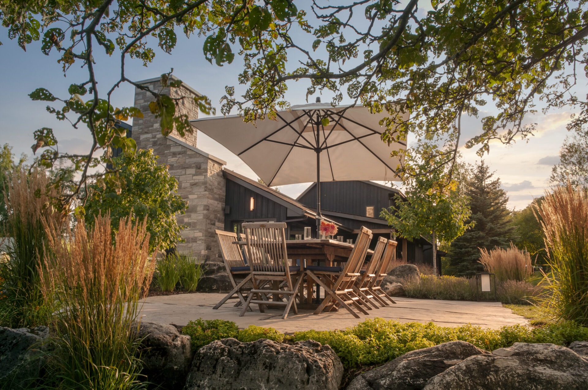 A serene outdoor patio features wooden furniture, large umbrella, and lush greenery, framed by leafy trees and a stone chimney backdrop.