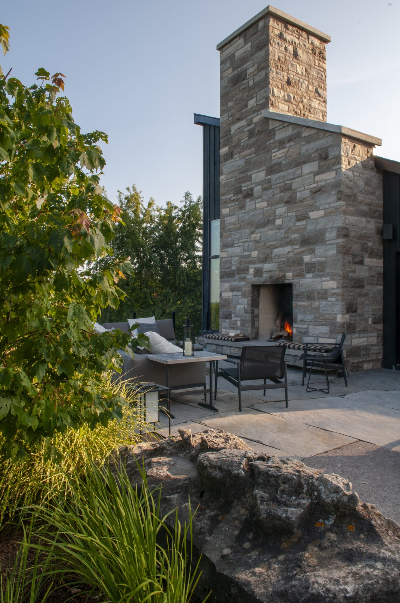 Modern outdoor patio with stone fireplace, surrounded by greenery. Comfortable seating area with chairs, a table, and a small fire burning.