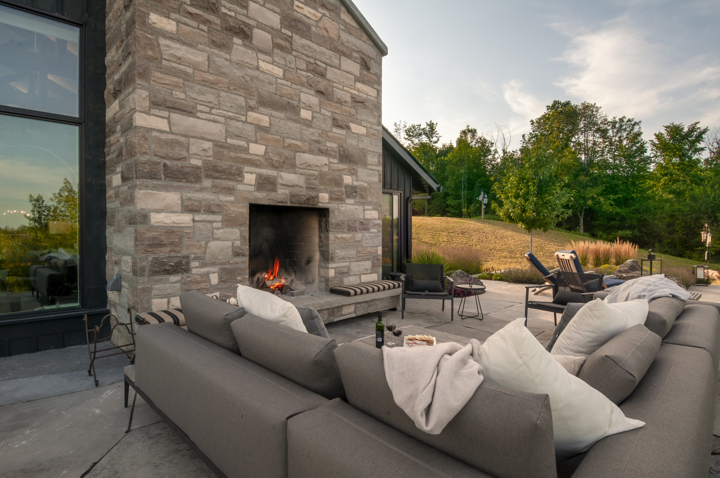 Cozy outdoor patio with stone fireplace, comfortable seating, wine glasses, and lush greenery in the background under a clear sky.
